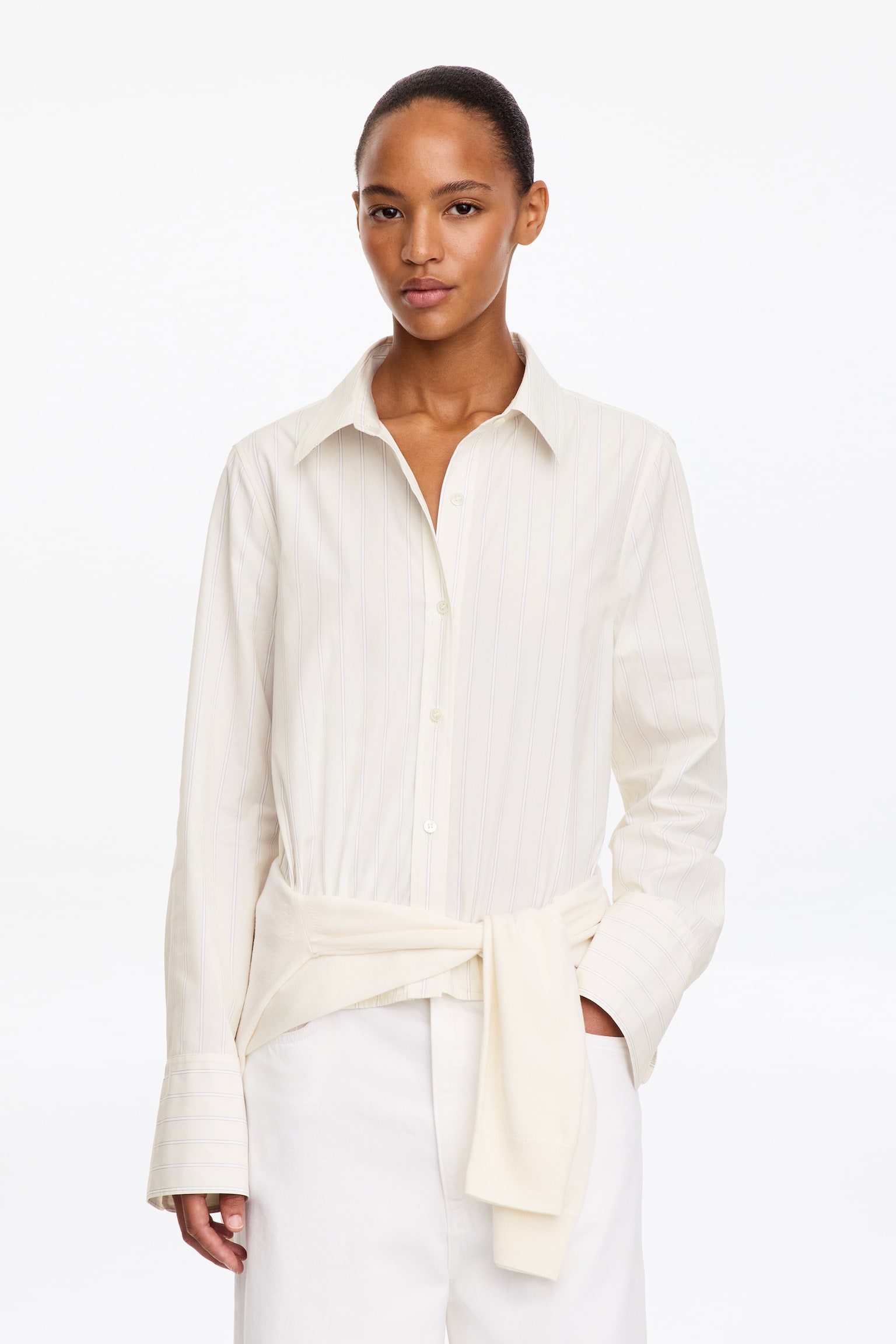 La modella indossa una camicia bianca in stile smoking a righe sottili, con colletto a punta e polsini ampi. Un maglione color crema è annodato in vita sopra la camicia, abbinata a pantaloni bianchi. - 1