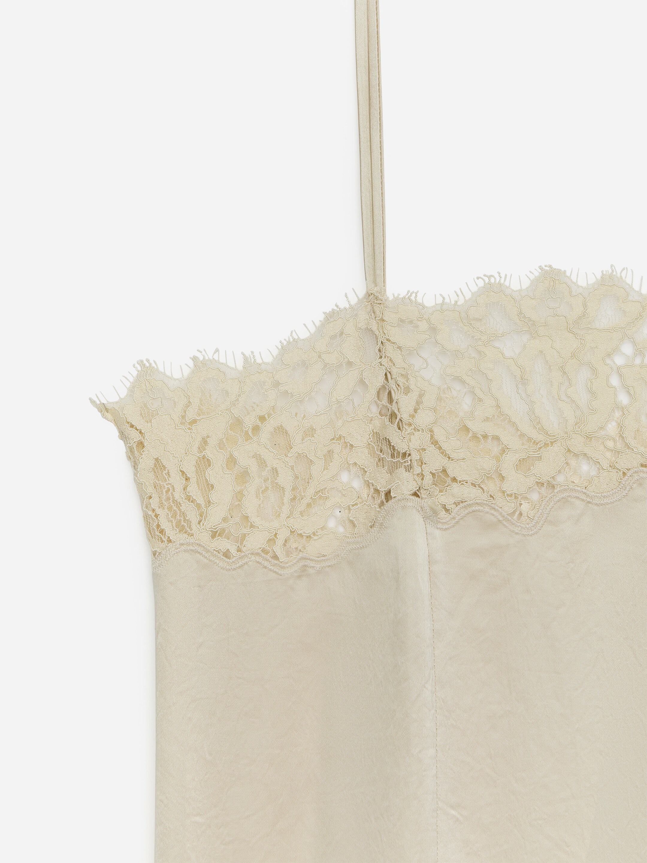 Agrandir l'image: Un caraco crème met en valeur une bordure en dentelle complexe le long de son bord supérieur, mettant en vedette un design festonné. Une fine bretelle s'étend vers le haut, et le corps principal du vêtement présente une texture légèrement froissée.