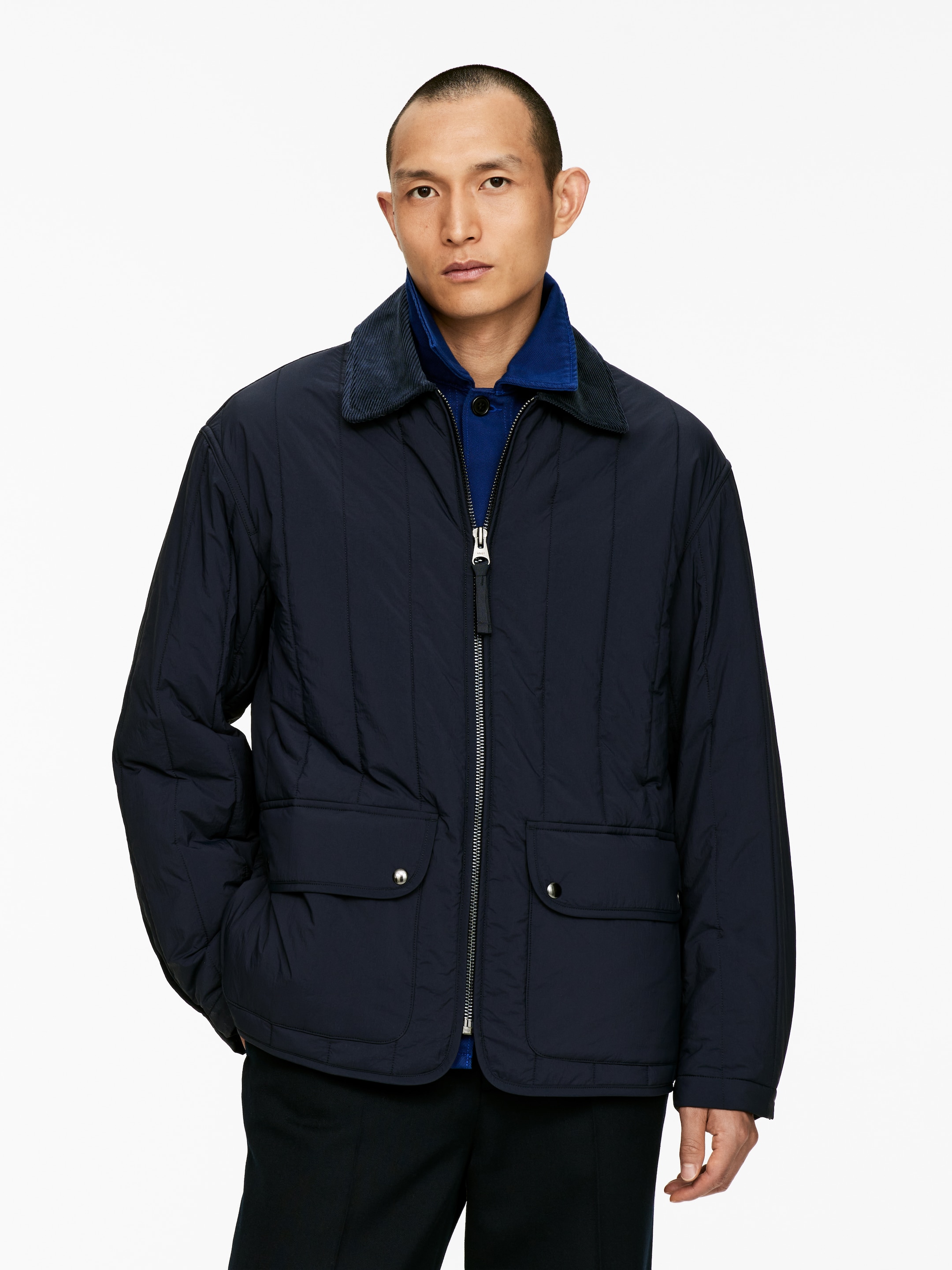 Grösseres Bild ansehen: Eine marineblaue gesteppte Jacke wird getragen, die einen kontrastierenden dunklen Cordkragen, einen silberfarbenen Reißverschluss in voller Länge und zwei große Klappentaschen mit Druckknopfverschlüssen an der unteren Vorderseite aufweist. Sie ist über einem leuchtend blauen Hemd geschichtet.