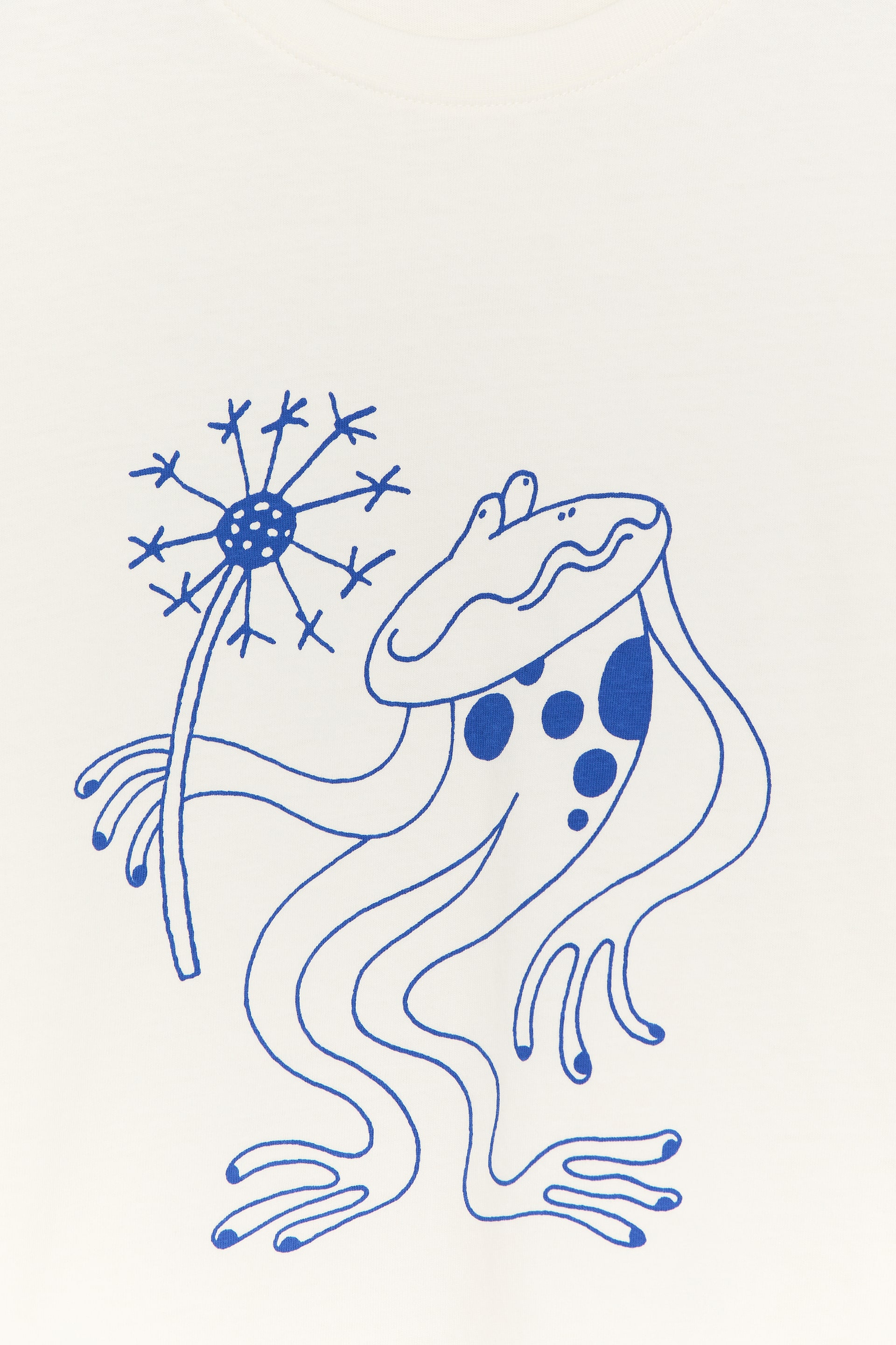 Ver imagem maior: A frente de uma t-shirt de cor creme exibe um desenho de linha azul de um sapo, que está segurando um dente-de-leão. O corpo do sapo apresenta bolinhas azuis.