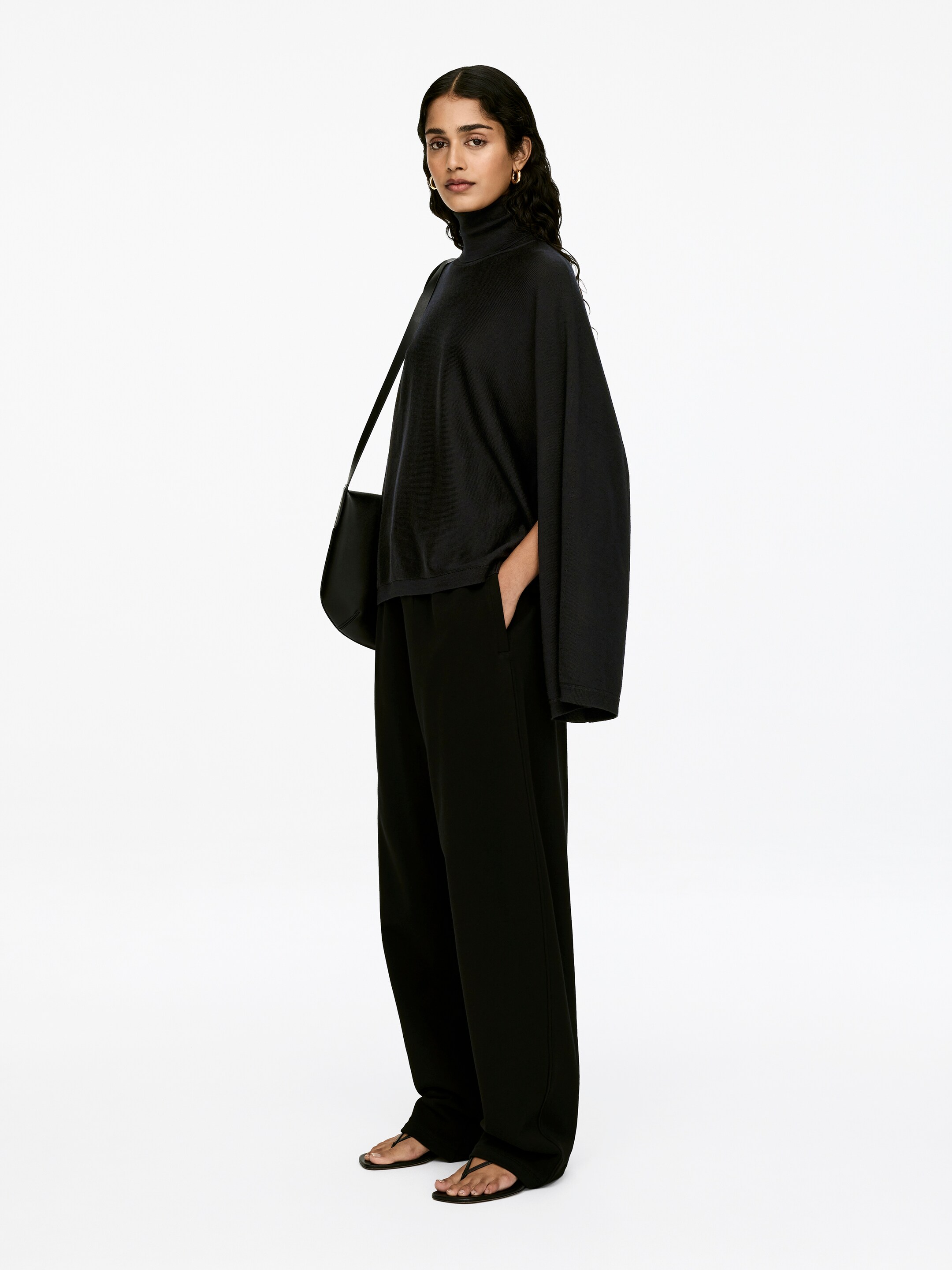 Agrandir l'image: Le mannequin porte un pull à col roulé noir avec une coupe ample et des bordures côtelées, qui présente des découpes aux emmanchures permettant à son bras droit d'être positionné à l'intérieur du pull, avec la main reposant dans une poche de pantalon.