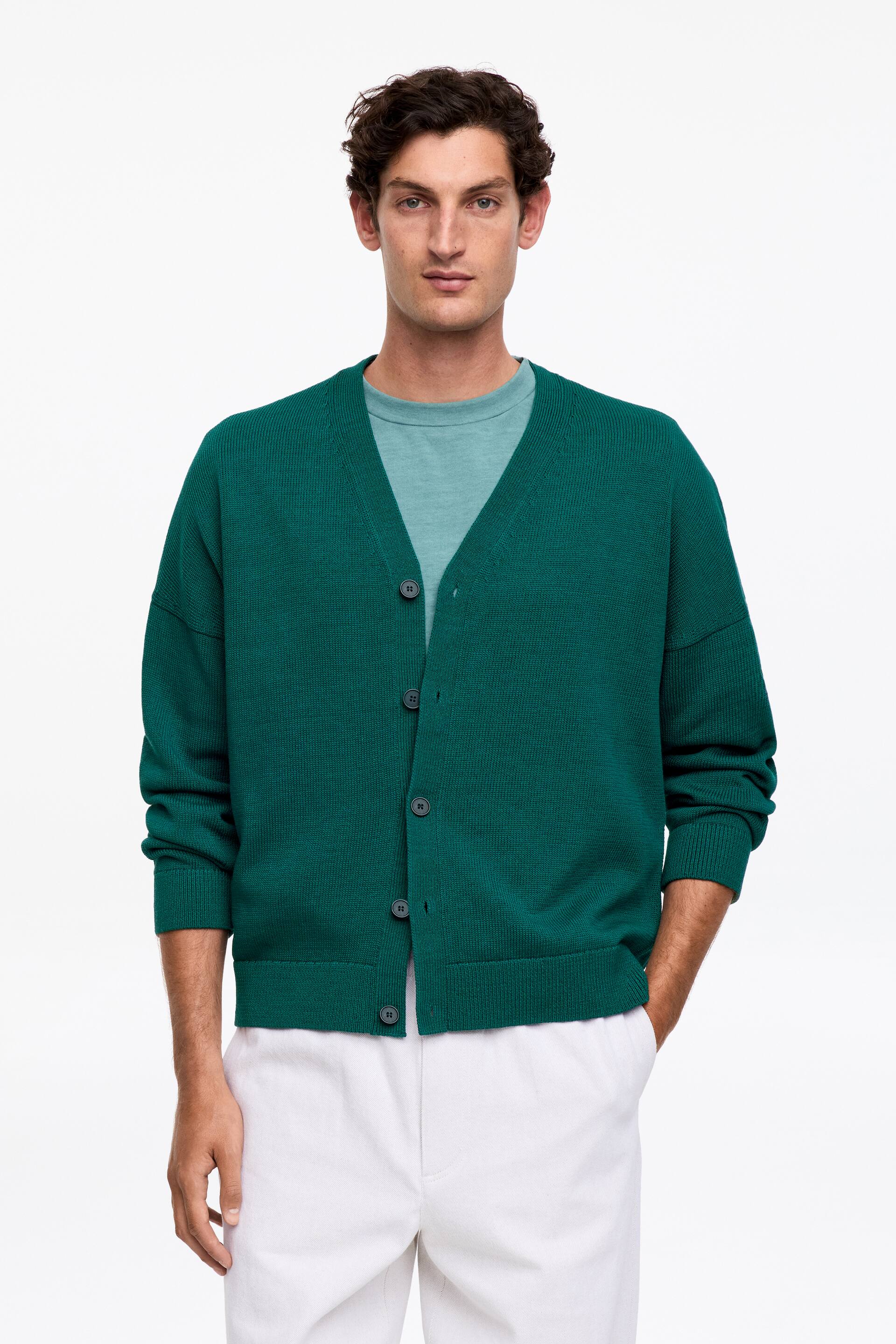 Προβολή μεγαλύτερης εικόνας: Σκούρο πράσινο ζακέτα V-λαιμόκοψη με κλείσιμο μπροστά με κουμπιά, πέντε σκούρα κουμπιά, μακριά μανίκια με ριγέ μανσέτες και ριγέ τελείωμα. Φοριέται ανοιχτή, πάνω από ένα ανοιχτό μπλε μπλουζάκι με στρογγυλή λαιμόκοψη.