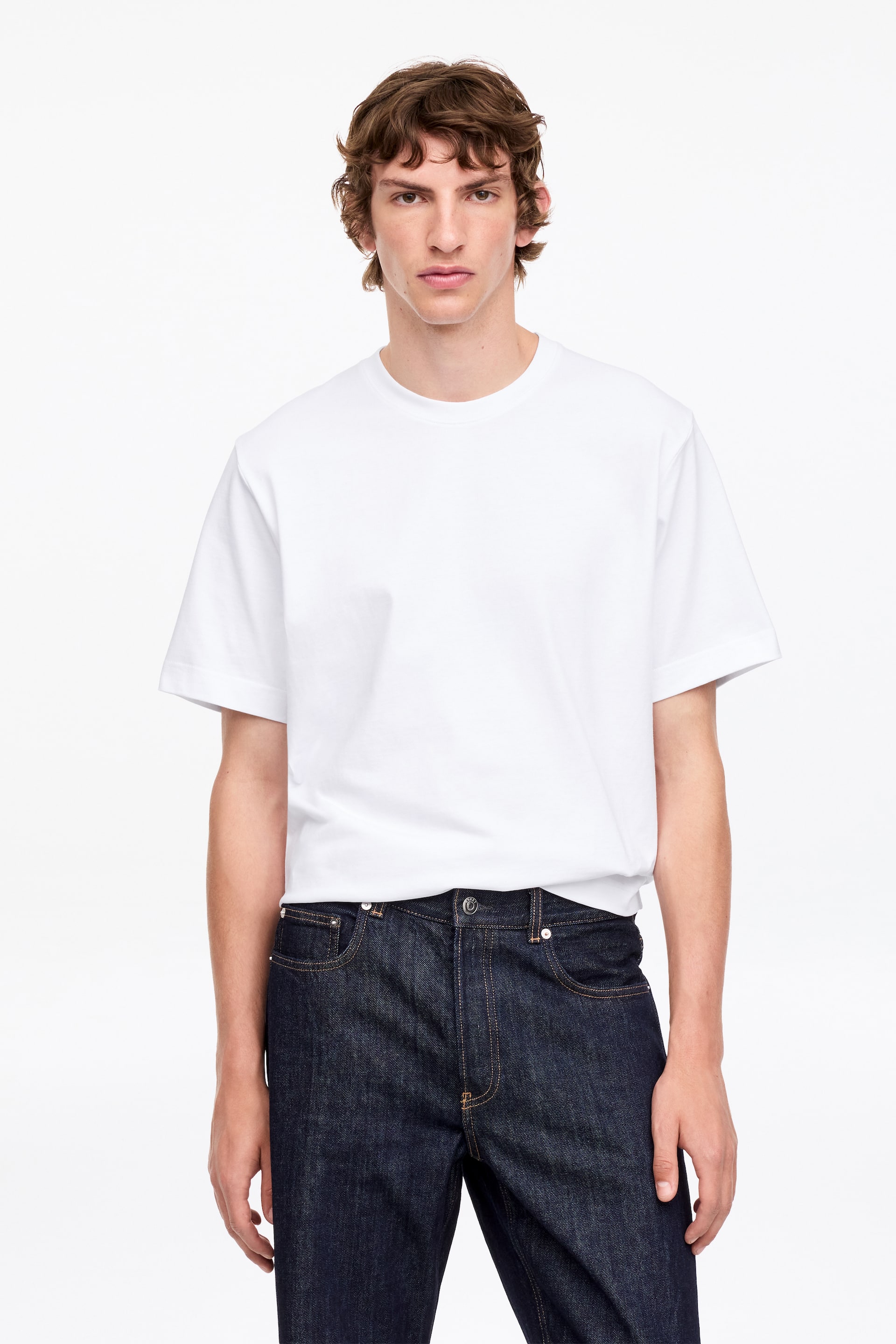 Ver imagen más grande: Una camiseta blanca con cuello redondo y mangas cortas de ajuste holgado, que presenta una silueta suelta y cuadrada. Su dobladillo está parcialmente metido en unos vaqueros de lavado oscuro.