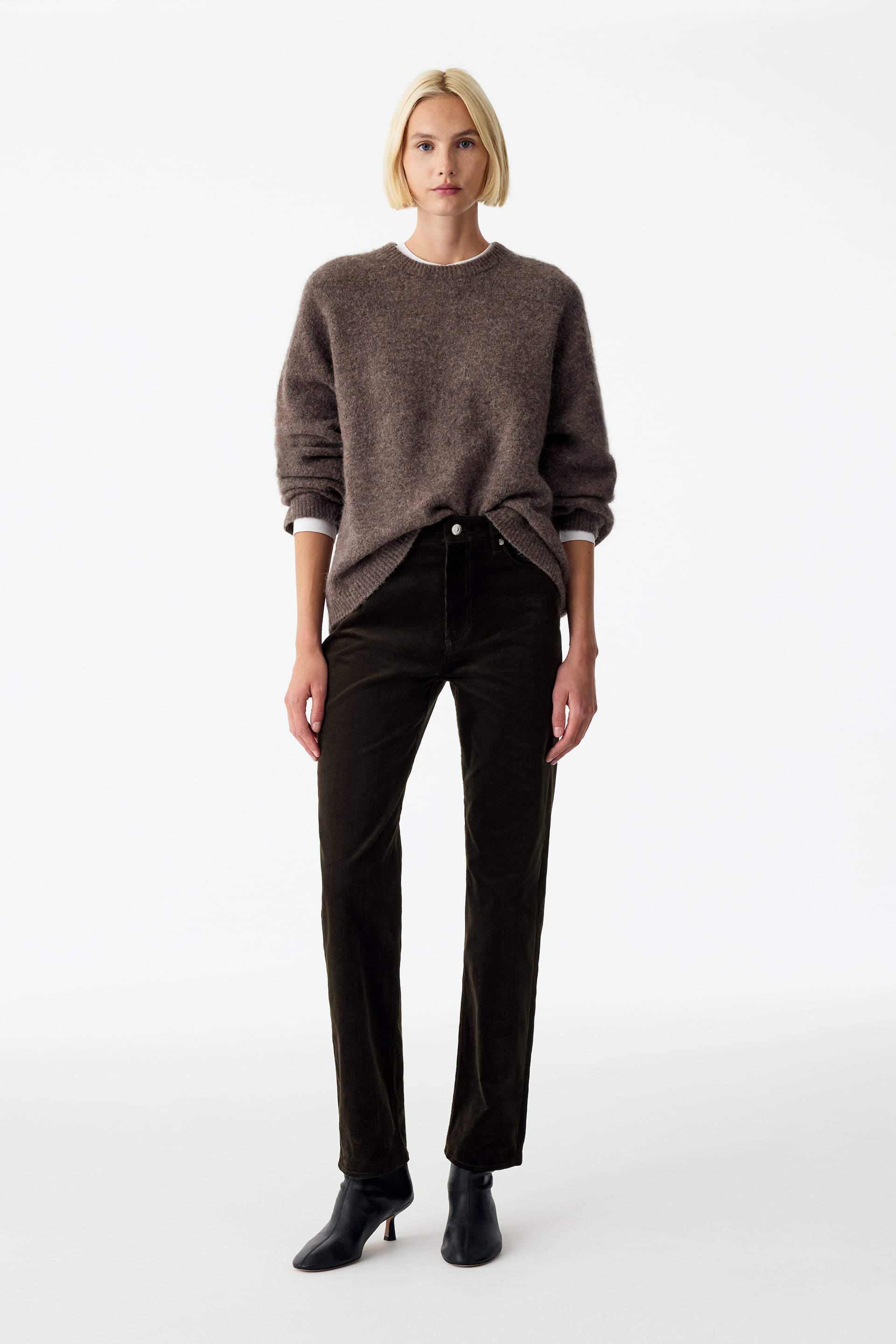 Visualizza immagine più grande: Pantaloni in velluto a coste marrone scuro, slim fit, con gamba dritta leggermente corta, indossati dalla modella. I pantaloni sono abbinati a un maglione lavorato a maglia marrone infilato nella parte anteriore in vita e a stivaletti neri con tacco.
