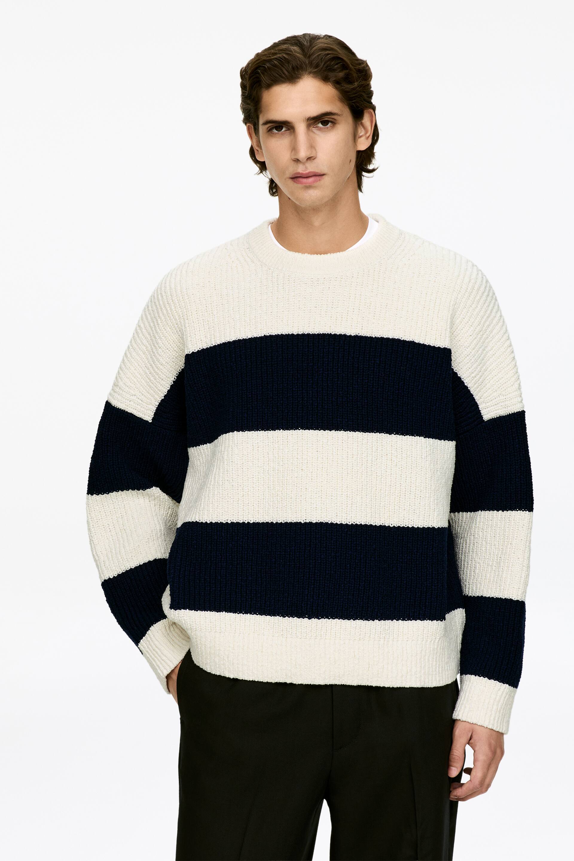 Vis større bilde: En krem- og marineblåstripet genser vender fremover, og viser en grovstrikket tekstur. Den har brede horisontale striper i vekslende krem og marineblå, en rund hals, lange ermer med ribbestrikkede mansjetter og en ribbestrikket kant. En hvit t-skjorte er synlig under den runde halsen.