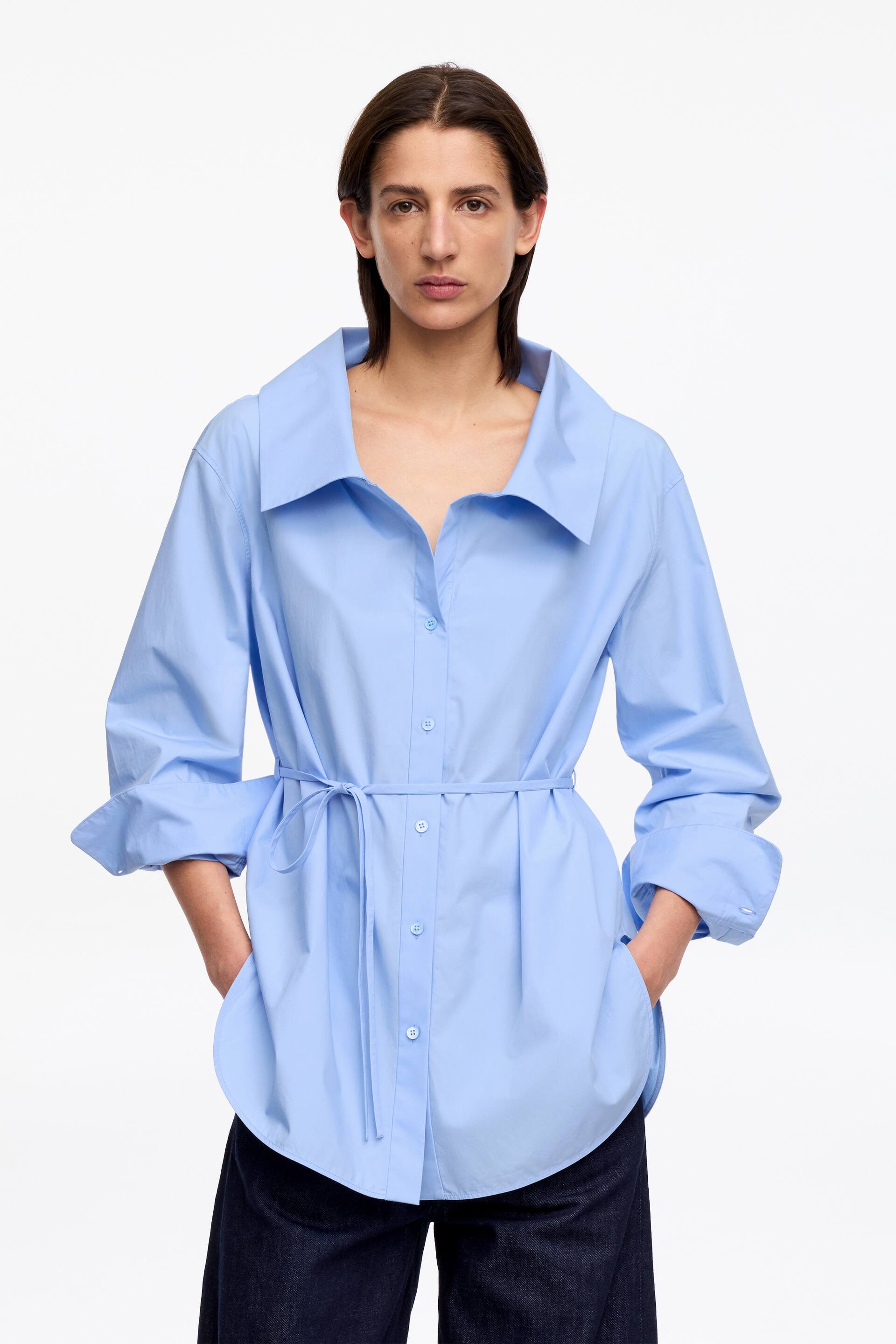Преглед на по-голямо изображение: Светлосиня широка риза е представена с лице напред, отличаваща се с широка, свободна яка и закопчаване с копчета отпред. Дългите ръкави са навити, а колан от същата материя стяга талията, създавайки меки гънки над закръглен подгъв на ризата.