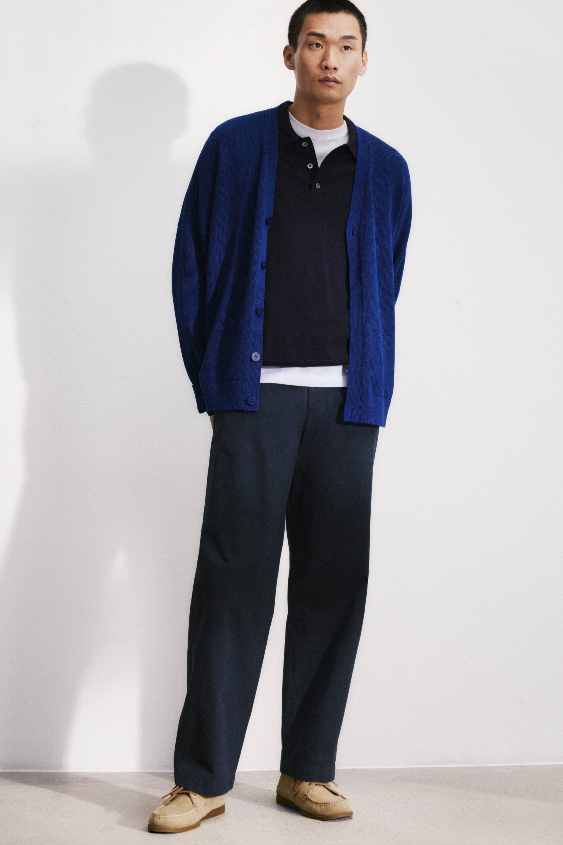 Se større billede: En blå cardigan med åben front har en afslappet pasform med runde knapper langs stolpen og ribbede manchetter, der lagres over en mørk marineblå kortærmet poloshirt. Poloshirten har en tre-knaps stolpe og klassisk krave, og afslører en hvid undertrøje ved kanten. Mørk marineblå bukser med brede ben bæres med lysebrune snørede mokkasin-stil sko.