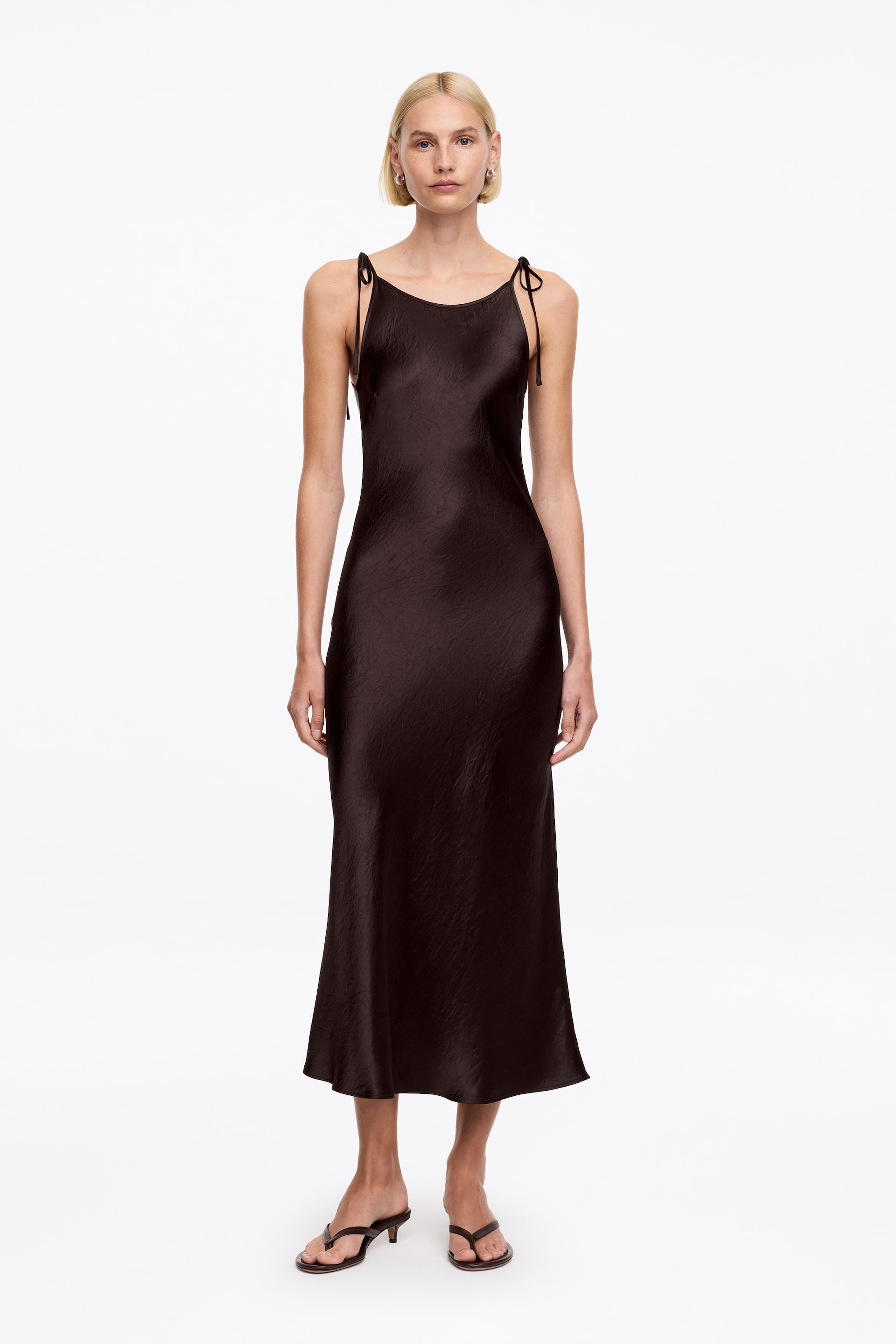 Näytä suurempi kuva: Tummanruskea slip-mekko, jossa on pyöreä pääntie ja ohuet spagettiolkaimet, jotka on solmittu olkapäille. Mekko on suunniteltu midi-pituiseksi ja siinä on teksturoitu pinta, joka luo kapean siluetin edestäpäin.
