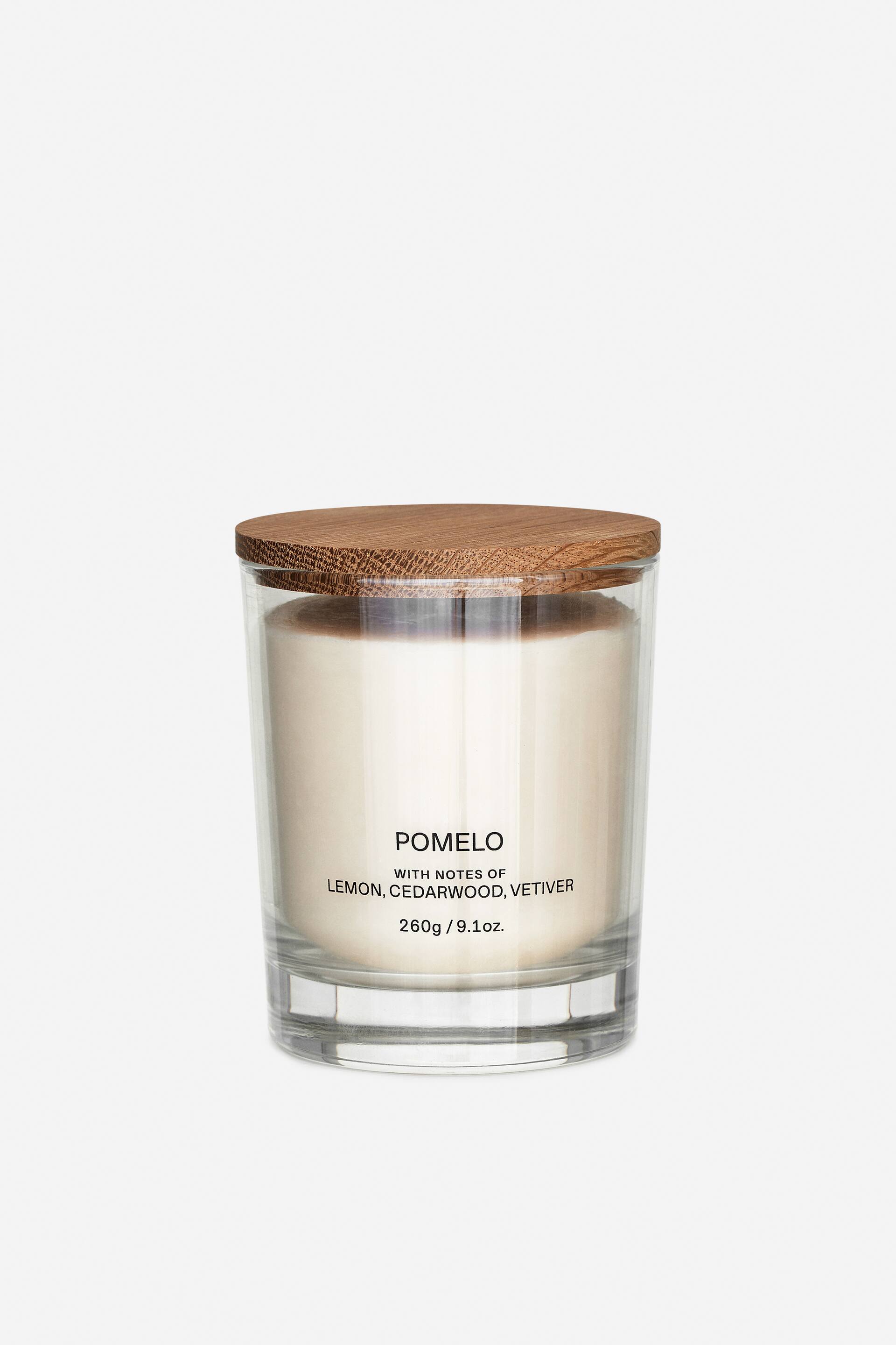 Agrandir l'image: Une bougie cylindrique est placée à l’avant, enfermée dans un récipient en verre transparent et surmontée d’un couvercle rond en bois. Une cire de couleur claire remplit le contenant, et le texte sur le verre indique « POMELO with notes of lemon, cedarwood, vetiver 260g / 9.1oz. »