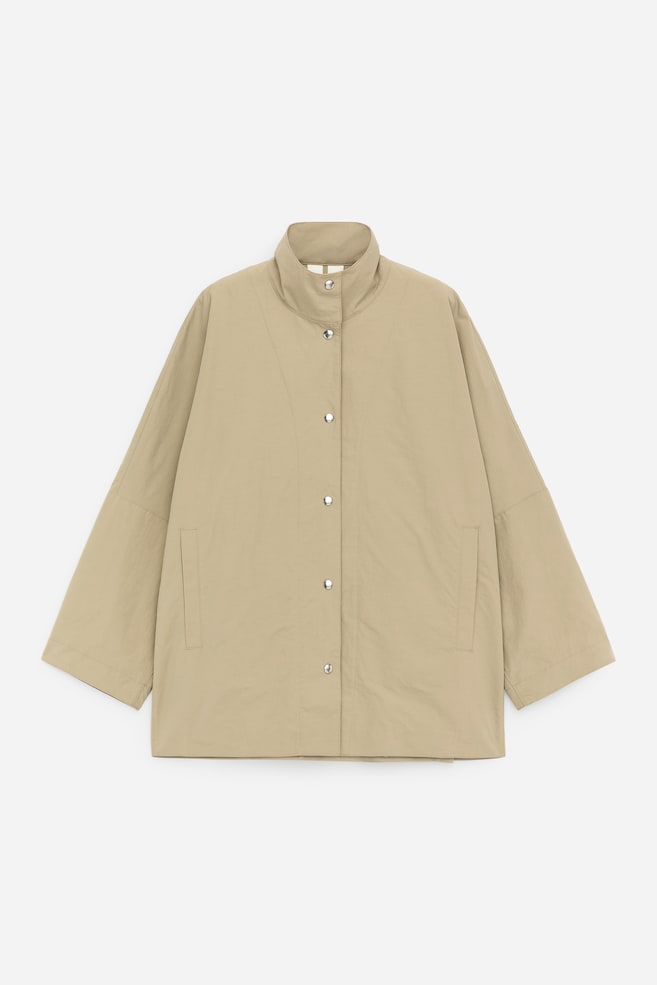 Eine beige Jacke ist von vorne zu sehen, mit einem Stehkragen und einer durchgehenden Knopfleiste, die mit silberfarbenen Druckknöpfen gesichert ist. Sie verfügt über weite, dreiviertellange Ärmel und zwei vertikale Schlitz-Taschen auf der Vorderseite. - 1