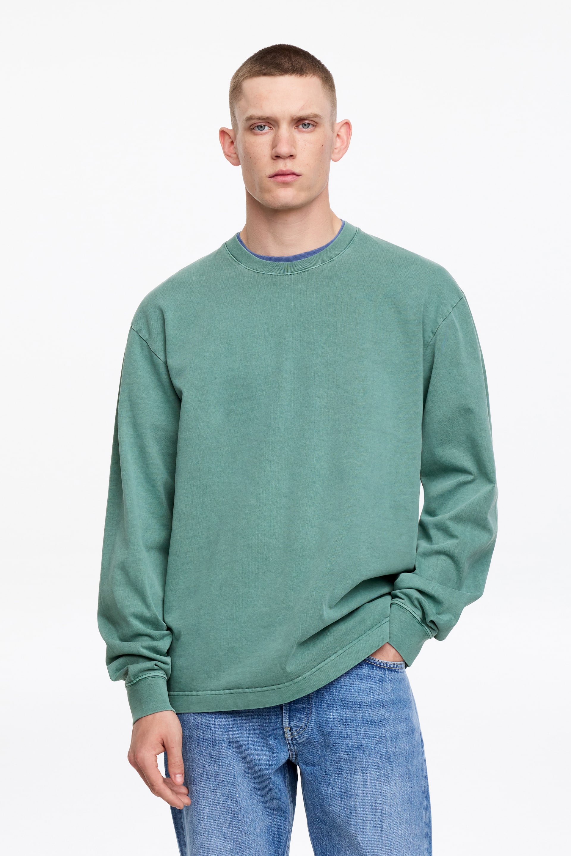 Zobrazit větší obrázek: Na sobě je vypraný zelený crewneck svetr, který je otočený dopředu a má dlouhé rukávy s žebrovanými manžetami a žebrovaným lemem. Zpod výstřihu vykukuje modré crewneck tričko.
