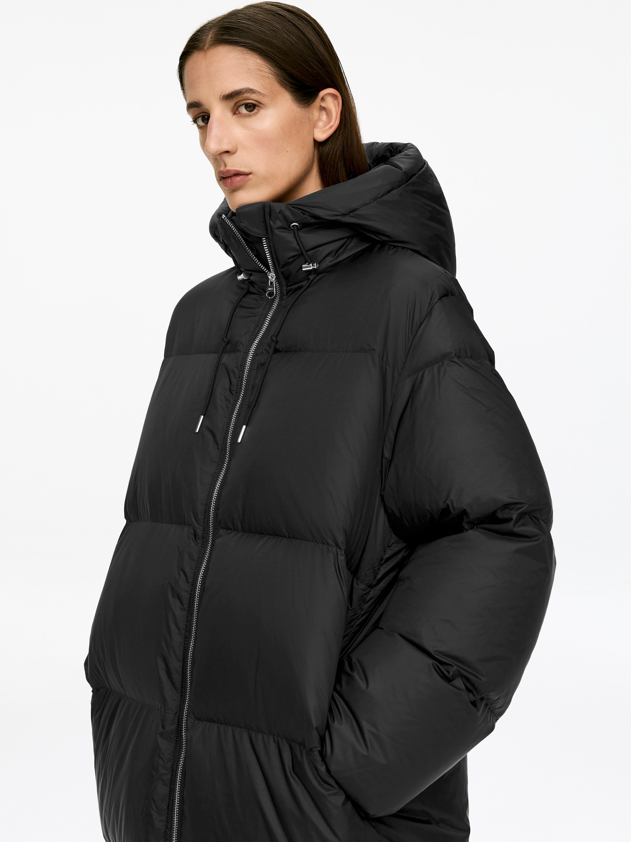 View larger image: An Upcycled Down Long Coat in black is being worn by the model, featuring a visible silver-toned zipper down the front and an attached hood that is up.