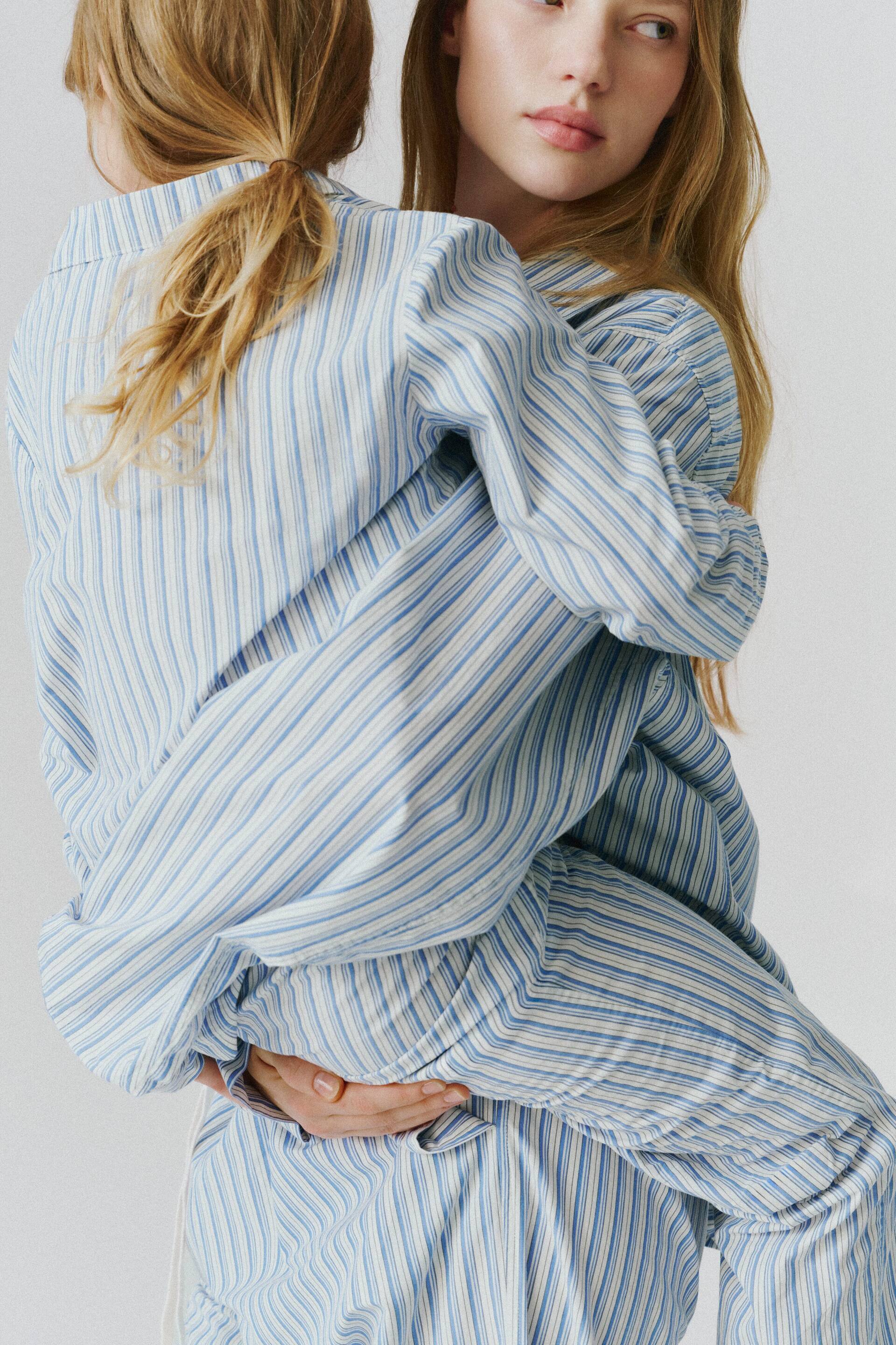 View larger image: A matching blue and white striped cotton pyjama set is being worn by two individuals, showcasing the relaxed-fit, long-sleeved shirt and matching trousers. The garments feature a classic vertical stripe pattern in light blue and white.