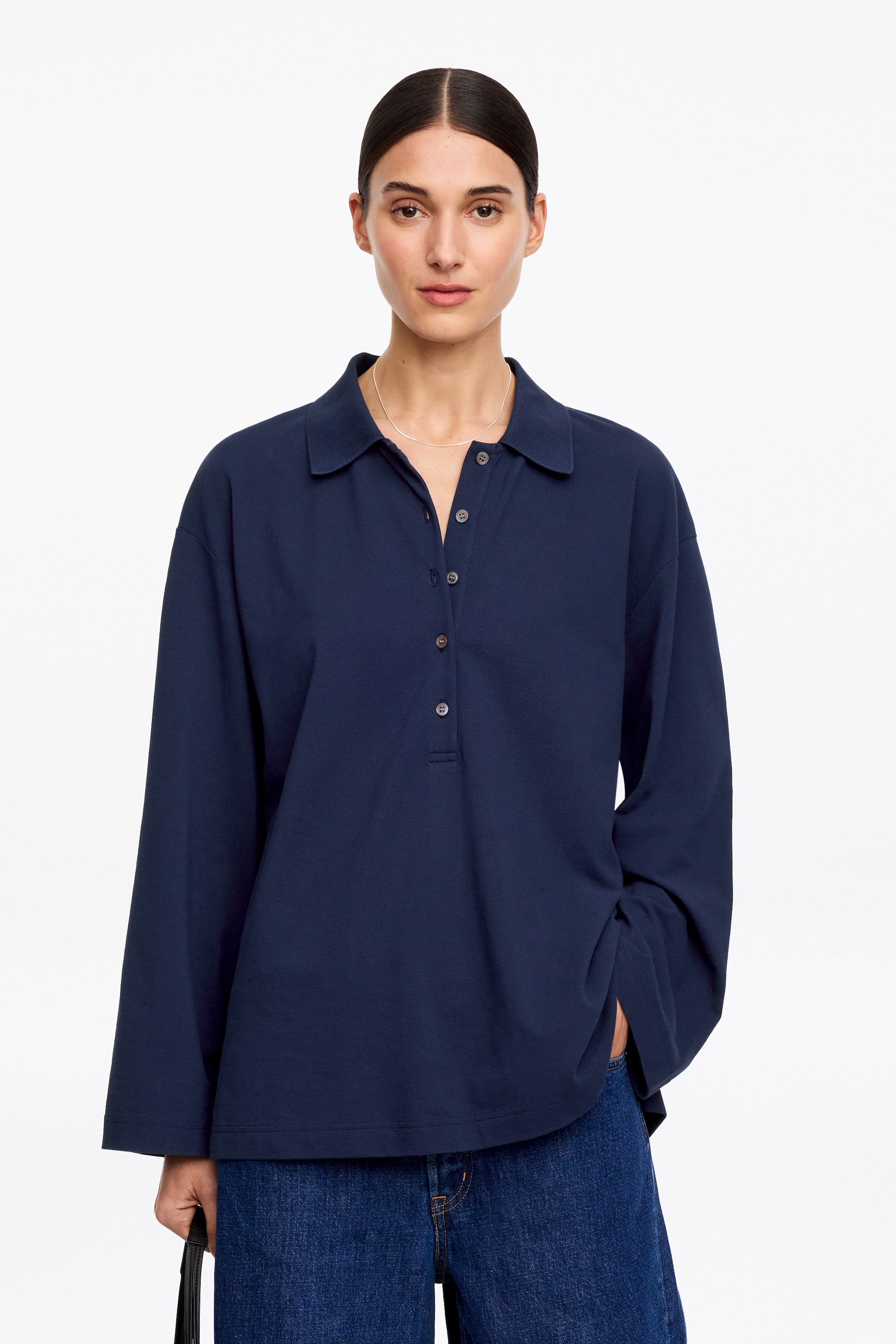 Προβολή μεγαλύτερης εικόνας: Η μοντέλο φοράει ένα σκούρο μπλε πικέ πόλο πουκάμισο. Διαθέτει γιακά, κουμπωτή πατιλέτα και μακριά, φαρδιά μανίκια. Ένα ασημένιο κολιέ βρίσκεται πάνω στο γιακά του πουκαμίσου, και το ένα χέρι της μοντέλου είναι τοποθετημένο μέσα στην τσέπη του παντελονιού, με το μανίκι του πουκαμίσου να πέφτει πάνω του.