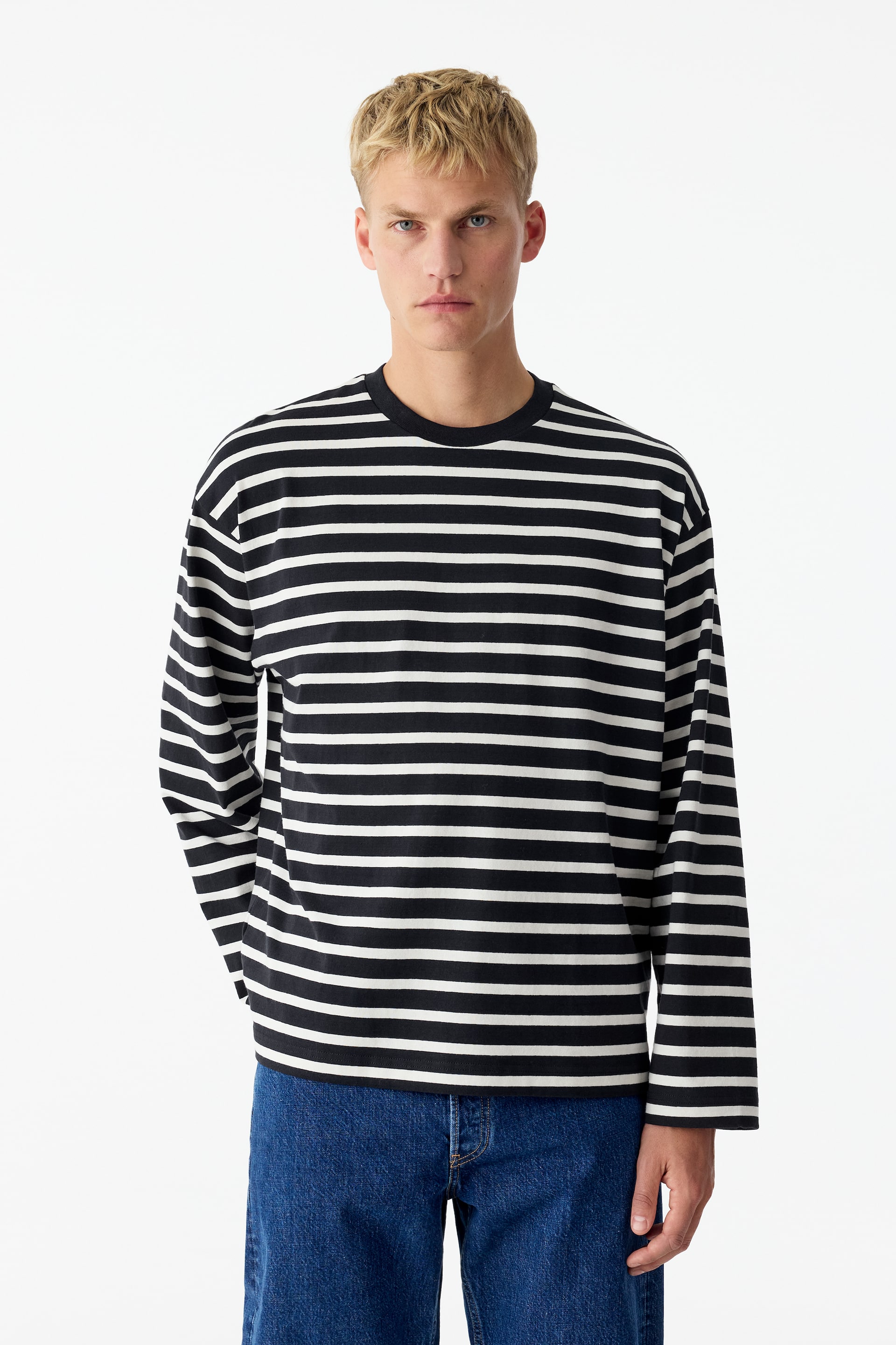 Se større billede: En T-shirt med afslappet pasform, lange ærmer og rund hals med sorte og hvide vandrette striber bæres af modellen. Modellen kombinerer T-shirten med blå jeans, mens hun står og vender fronten mod kameraet.
