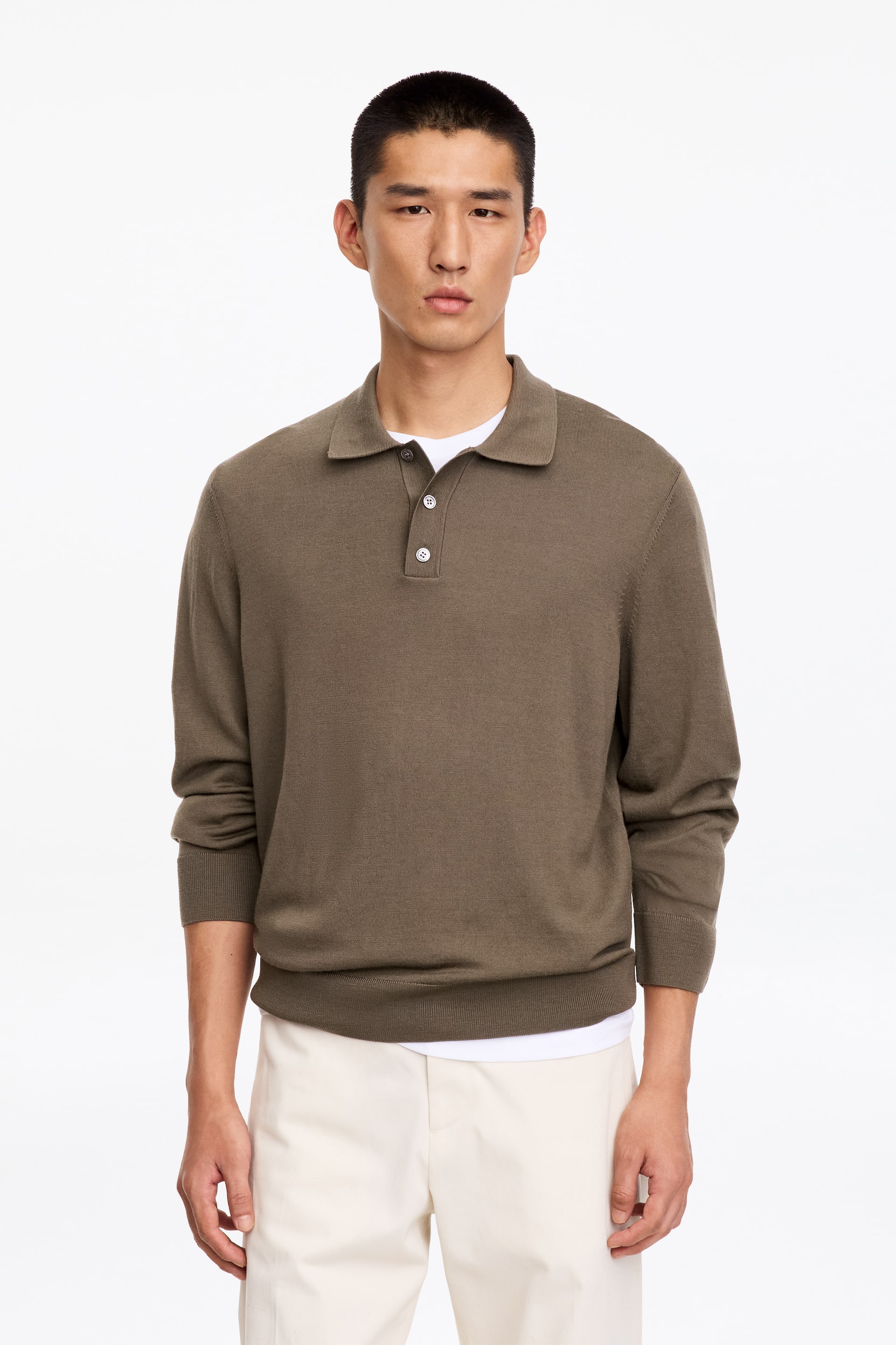 Agrandir l'image: Un pull polo en maille à manches longues, dans un brun sourd, est porté par le mannequin, avec un col classique, une patte de boutonnage à trois boutons et des finitions côtelées. Le pull est porté sur un t-shirt blanc et associé à un pantalon blanc cassé.