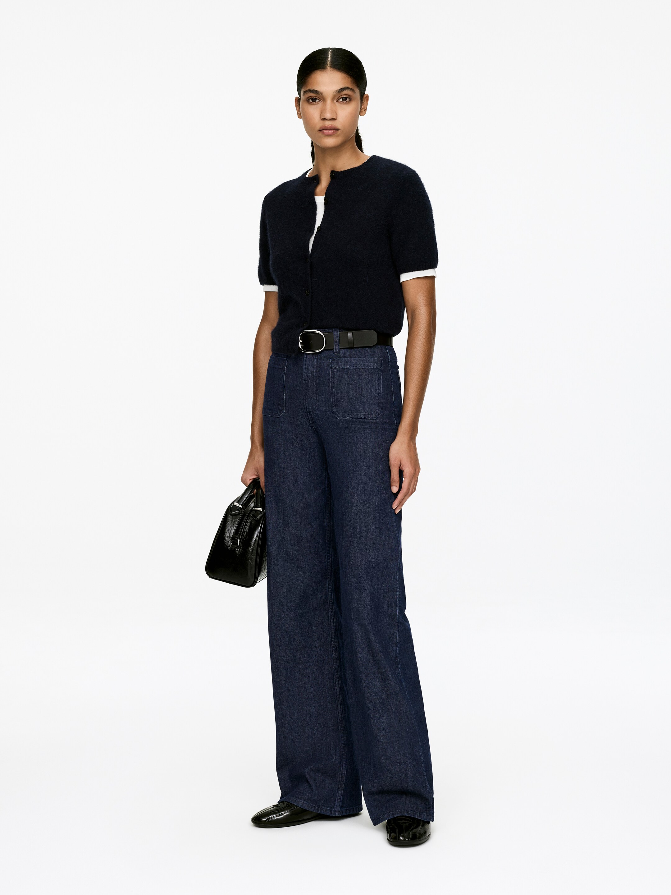 Agrandir l'image: Un ensemble orienté vers l'avant met en vedette un cardigan bleu marine à manches courtes et duveteux, superposé à un haut blanc à manches courtes, et un pantalon large indigo foncé avec des poches plaquées à l'avant. Une ceinture en cuir noir avec une boucle en argent serre la taille, des chaussures plates noires brillantes complètent le look, et un sac à main structuré noir est porté.