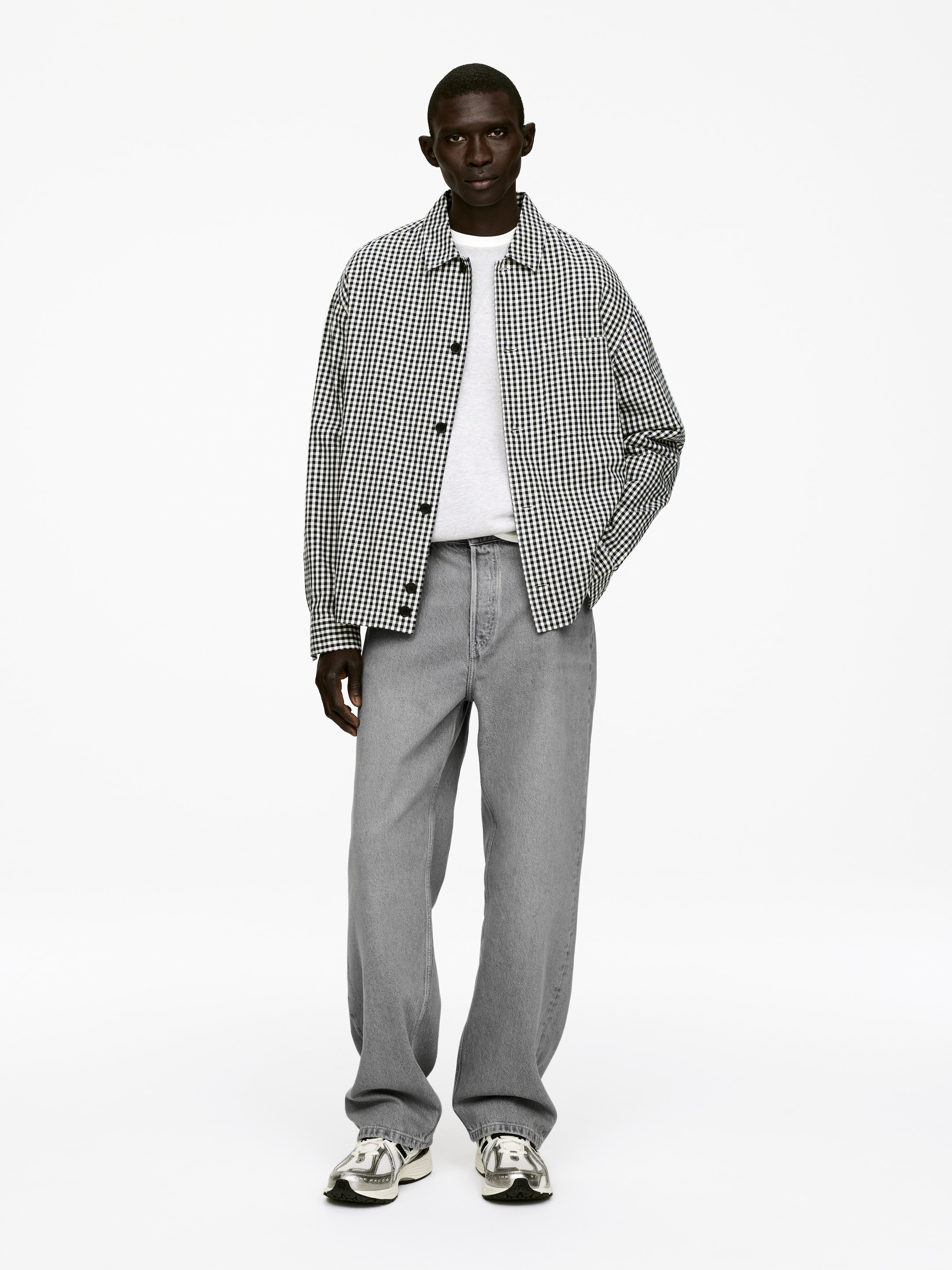 Ingrandisci l'immagine: Un outfit viene presentato, caratterizzato da una camicia giacca a quadri gingham bianca e nera con colletto aperto, indossata aperta su una t-shirt grigio chiaro con scollo a girocollo. Vengono indossati jeans grigi a gamba larga e vestibilità rilassata con scarpe sportive in argento metallizzato e nero.