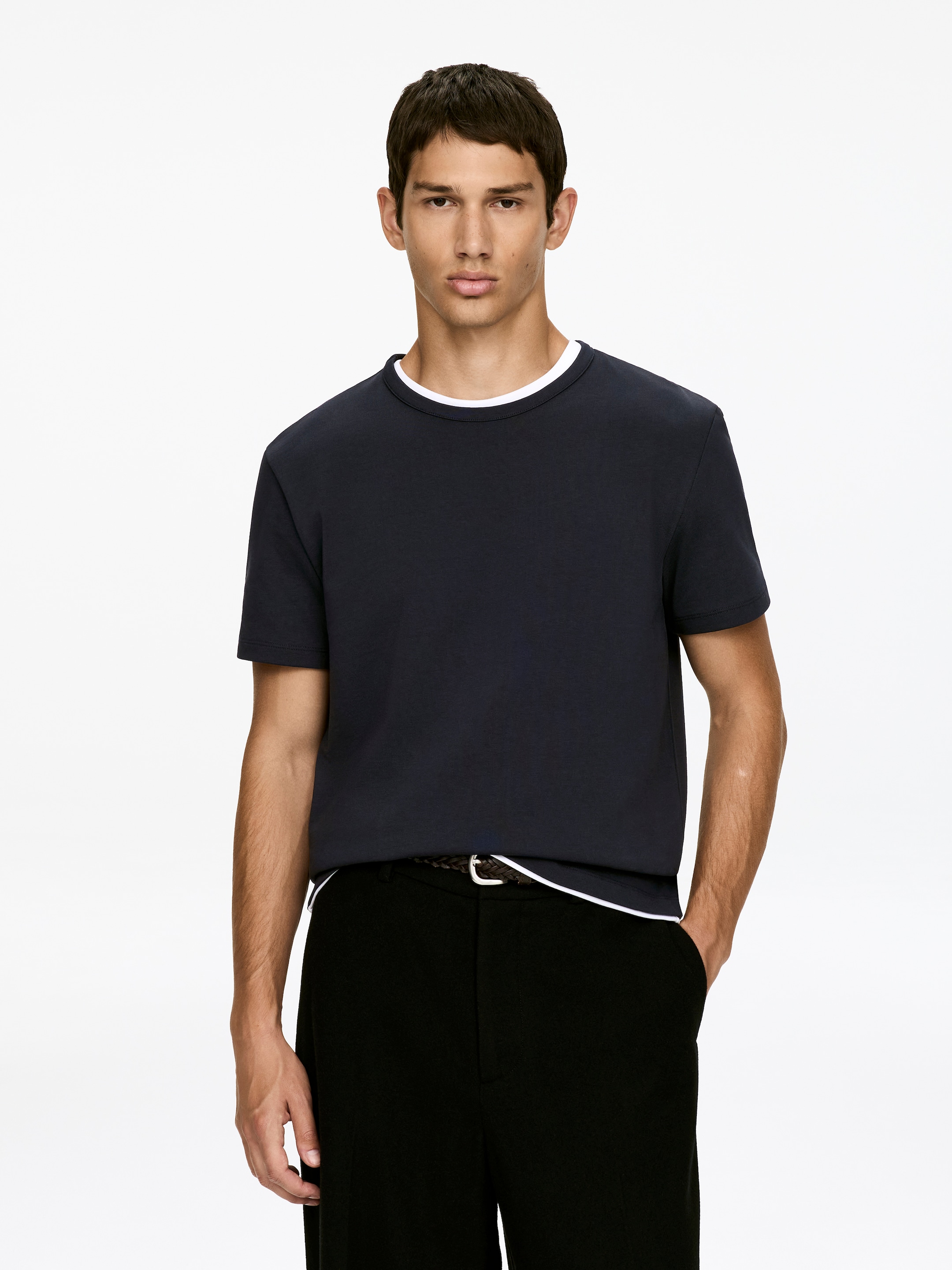 Visualizza immagine più grande: Una T-shirt girocollo blu navy, progettata con una vestibilità aderente sul petto e sulle braccia, è indossata dal modello. Uno strato bianco è visibile sotto la scollatura e l'orlo della T-shirt, indicando uno stile a strati.