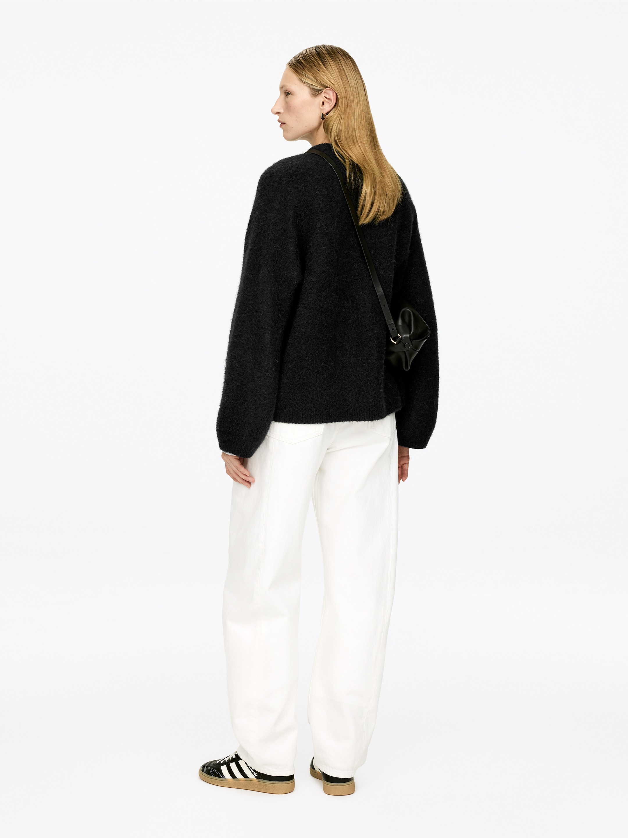 Grösseres Bild ansehen: Ein schwarzer Pullover mit texturierter Strickoptik wird aus einer Rückansicht in drei Vierteln präsentiert. Seine langen Ärmel enthüllen darunter weiße Manschetten, und eine kleine schwarze Crossbody-Tasche hängt über dem Pullover. Auch weiße Hosen mit weitem Bein werden gezeigt, kombiniert mit schwarz-weißen Sneakers, die Gummisohlen aufweisen.