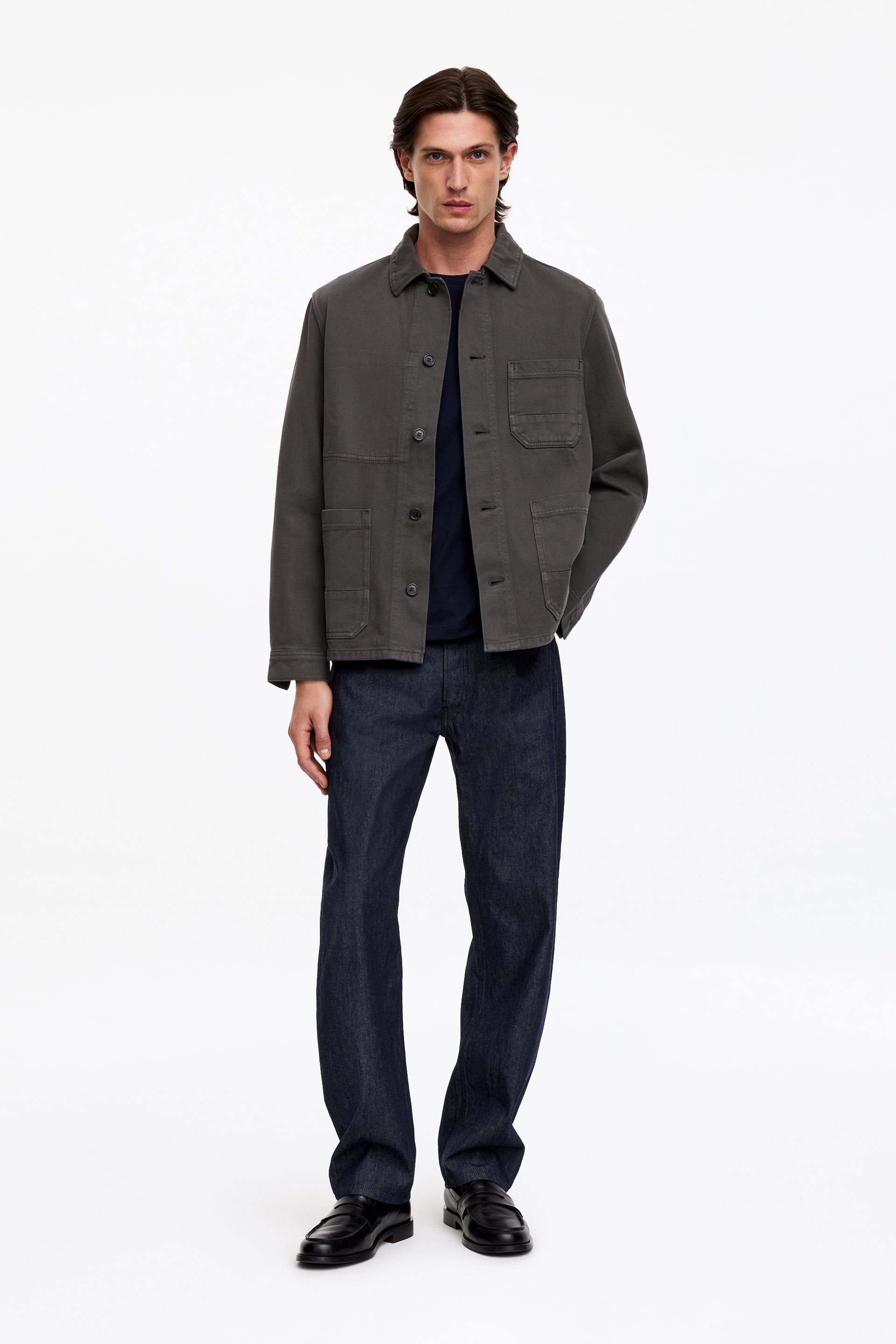 Ver imagen más grande: Una sobrecamisa de sarga de algodón en color gris oscuro y corte boxy la lleva la modelo desabrochada, superpuesta sobre una camiseta oscura de cuello redondo y unos vaqueros oscuros. La sobrecamisa presenta un cuello clásico, cierre delantero con botones, un único bolsillo de parche en el pecho y dos grandes bolsillos de parche en la parte delantera de la cadera.