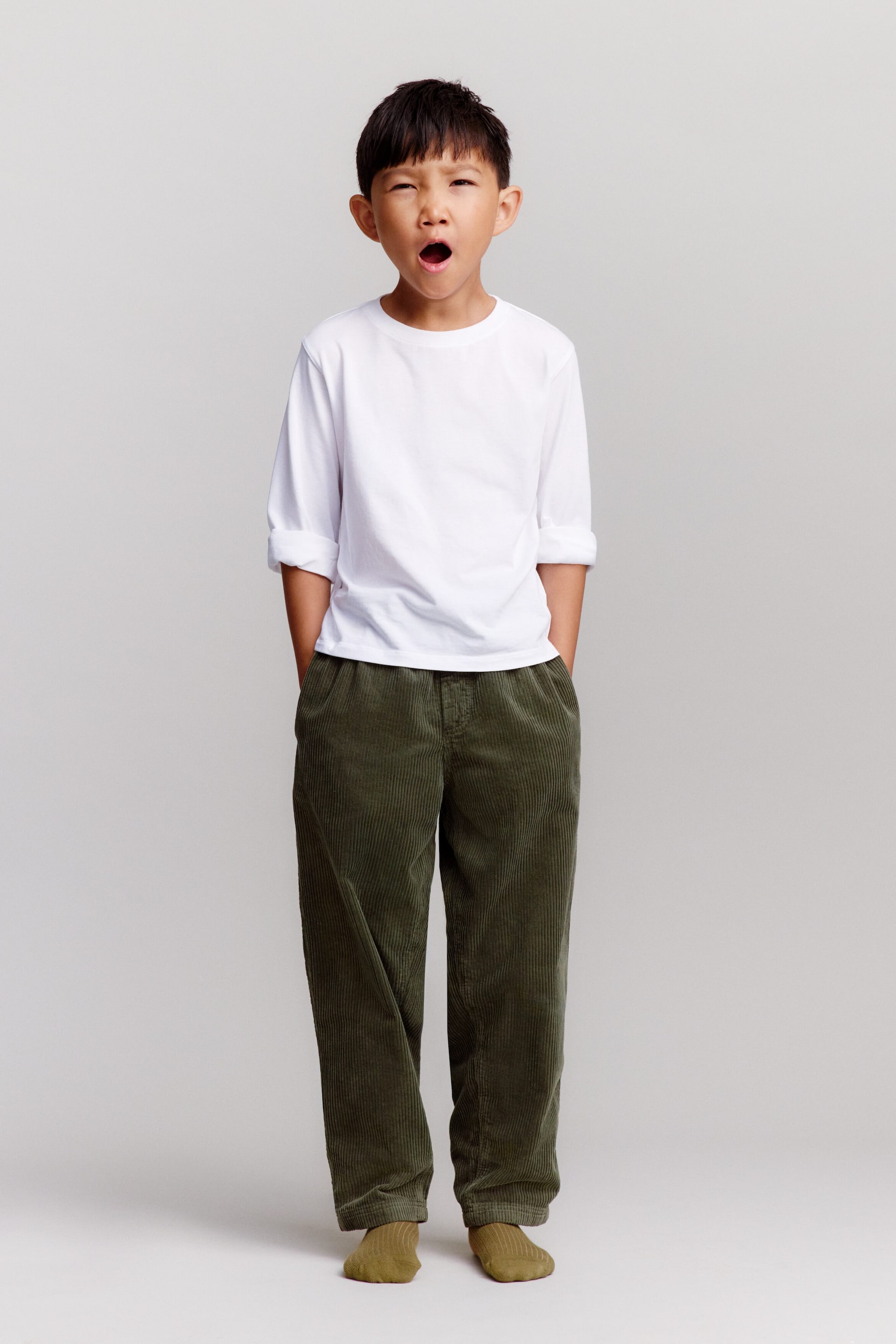 Visualizza immagine più grande: Un bambino indossa pantaloni in velluto a coste di cotone verde oliva dalla vestibilità comoda, con gamba affusolata e tasche laterali, mostrati frontalmente. I pantaloni sono abbinati a calzini a coste verde oliva coordinati e a una maglietta bianca a maniche lunghe.