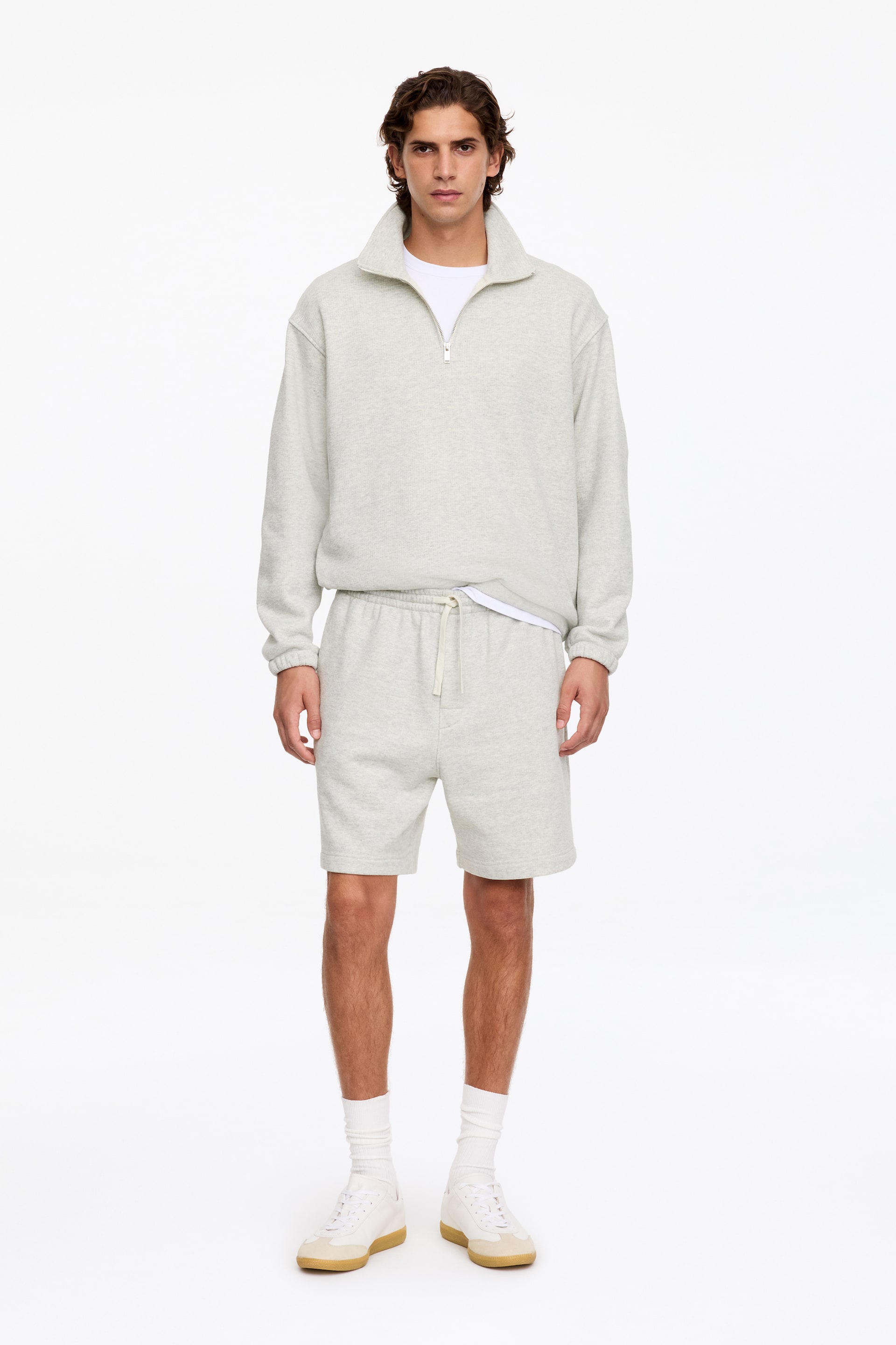 Größeres Bild ansehen: Lässig geschnittene Shorts aus leicht meliertem Jersey in Hellgrau werden vom Model getragen und zeichnen sich durch einen flachen Kordelzugbund sowie eine längere Silhouette aus. Die Shorts sind mit einem passenden Half-Zip-Loungewear-Sweatshirt, einem weißen T-Shirt, weißen Socken und weißen Sneakers kombiniert.