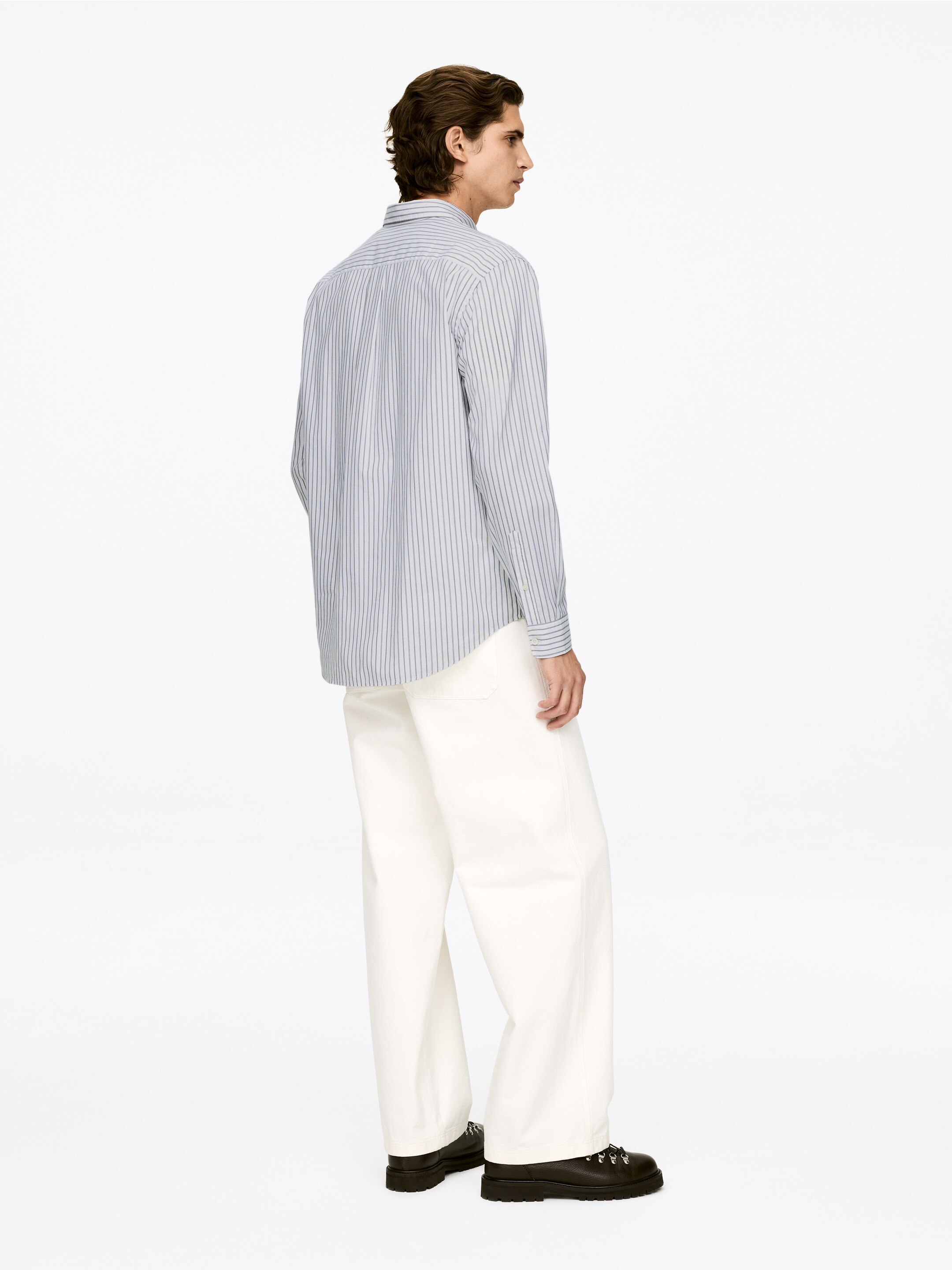 Visualizza immagine più grande: Una camicia con colletto e maniche lunghe con sottili righe verticali scure è presentata di lato e di spalle, indossata non infilata sopra pantaloni larghi color crema. Scarpe con suola spessa nere con lacci completano l'abbigliamento.