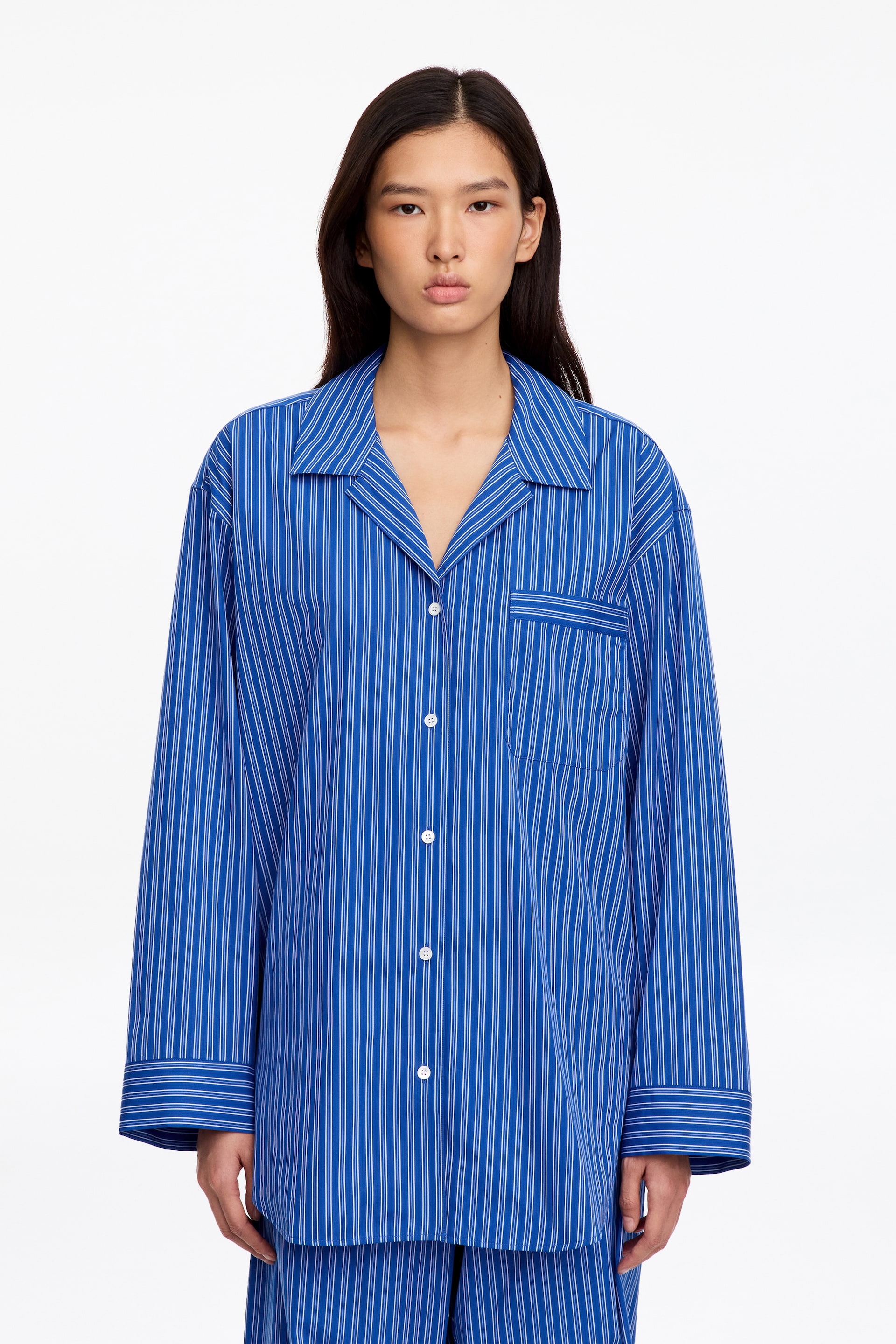 Ver imagem maior: A modelo veste uma camisa de pijama oversized às riscas azuis e brancas, com colarinho aberto e um bolso de chapa no peito. A camisa de manga comprida, com botões à frente, é combinada com umas calças de pijama às riscas a condizer.