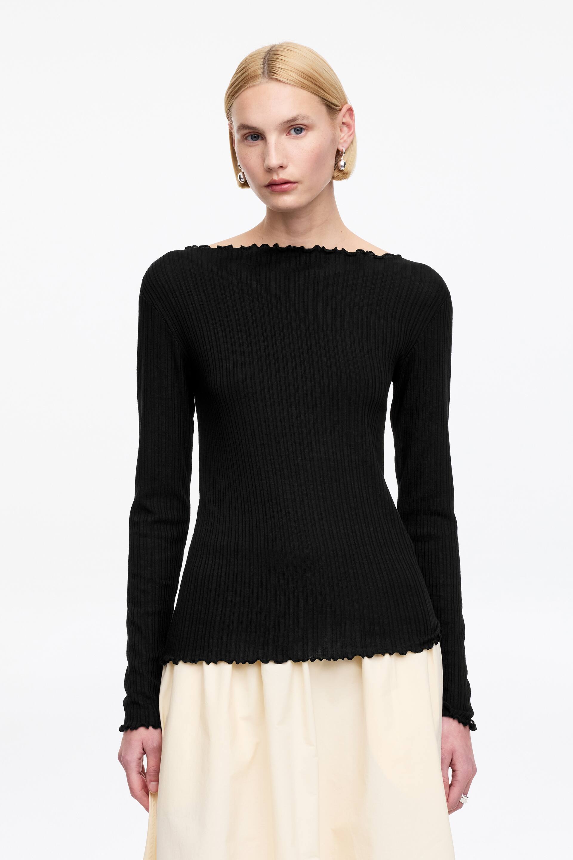 Visualizza immagine più grande: Un top nero a maglia a coste è mostrato frontalmente. Presenta uno scollo a barca, maniche lunghe e un orlo dritto, tutti rifiniti con bordo a lattuga. Il top aderisce perfettamente al corpo e termina all'altezza dei fianchi.