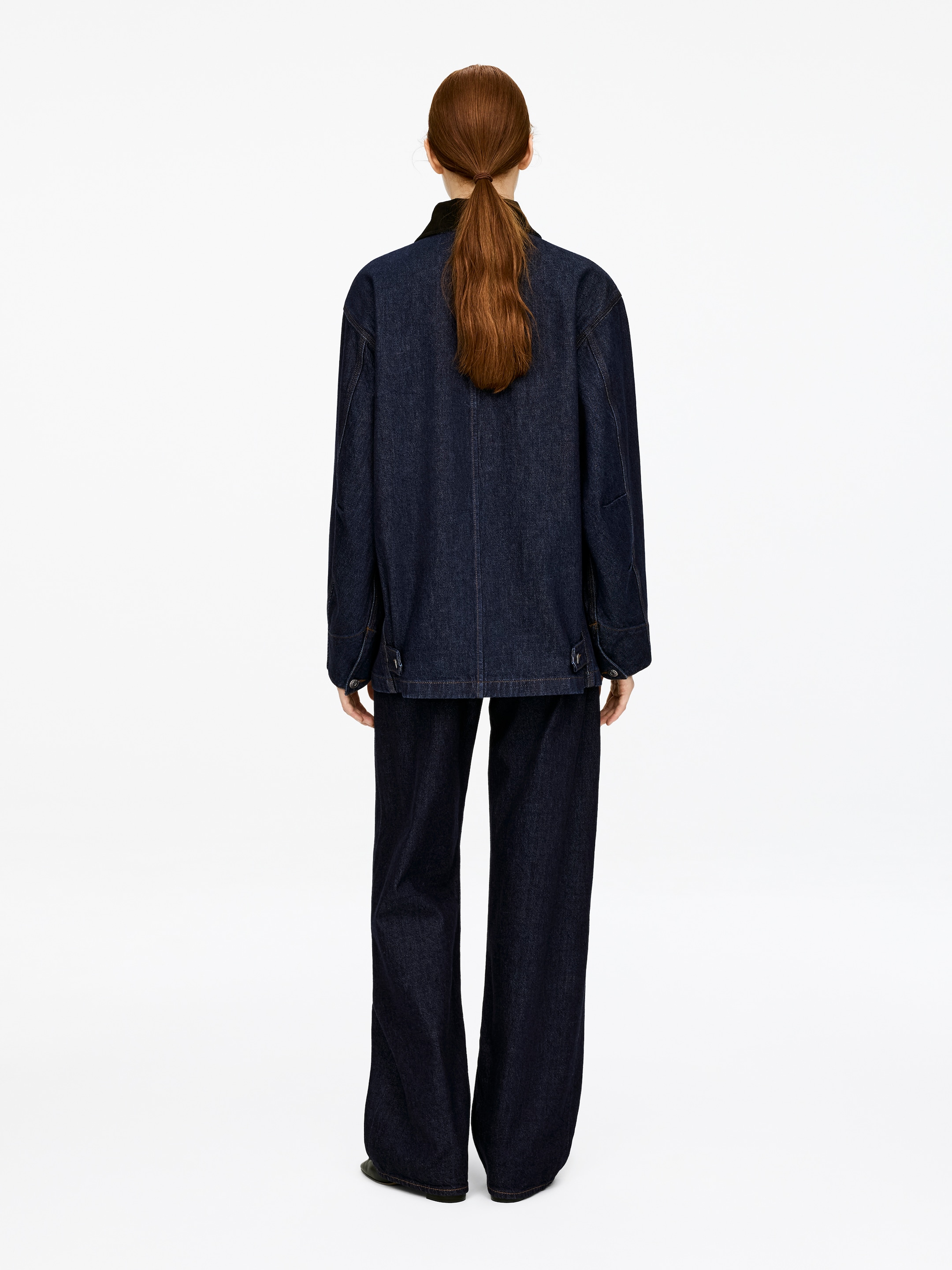 Visualizza immagine più grande: Una giacca di jeans scuro con un colletto in velluto a coste è indossata dalla modella. La giacca oversize presenta linguette laterali con bottoni a gambo in denim lungo l'orlo. La modella indossa anche dei pantaloni in denim scuro abbinati.