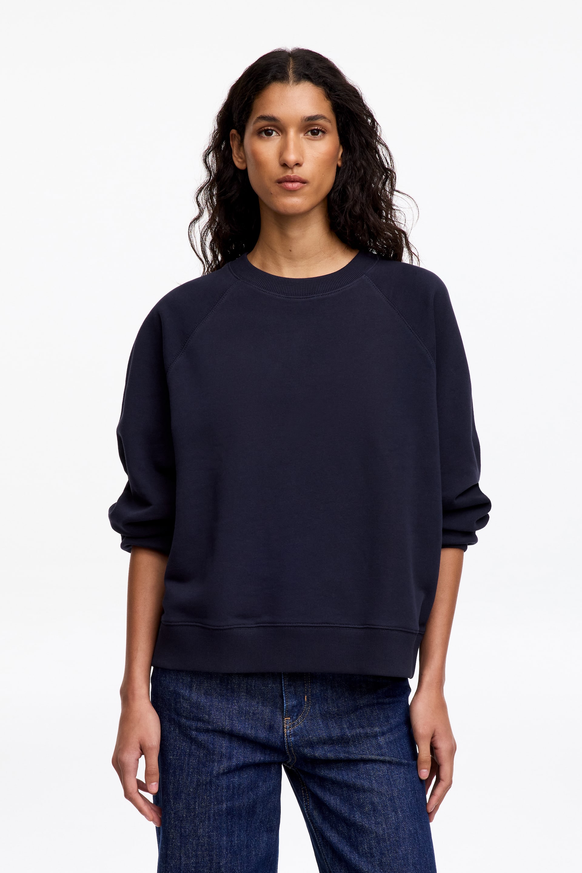 Grotere afbeelding bekijken: Het model draagt een donker marineblauwe sweatshirt met ronde hals, voorzien van raglanmouwen met opgestroopte boorden, een geribde kraag en een geribde zoom. De sweatshirt is gestyled met donkerblauwe jeans.