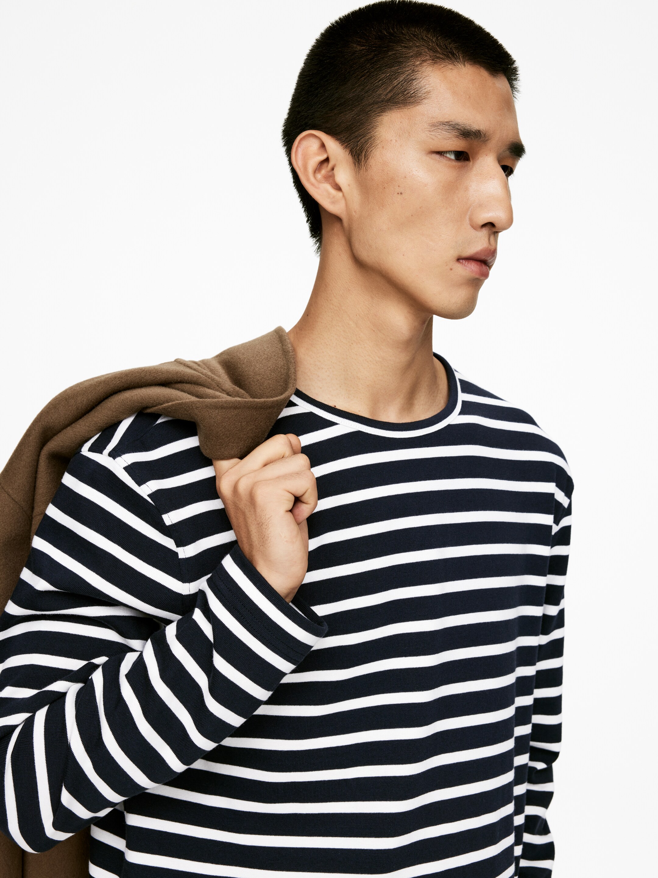 Grotere afbeelding bekijken: Een shirt met lange mouwen met horizontale marineblauwe en witte strepen, een ronde hals en geribde boorden wordt gedragen, gezien vanuit een zij-voorhoek. Een bruine jas wordt over de linkerschouder gedrapeerd.