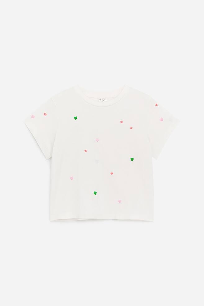Een gladde, crèmekleurige T-shirt toont zijn voorkant, ontworpen voor een wijde pasvorm met licht verlaagde schouders. Het is afgewerkt met een all-over borduurwerk van kleine roze, groene en rode hartvormen en heeft een geribbelde ronde halslijn. - 1