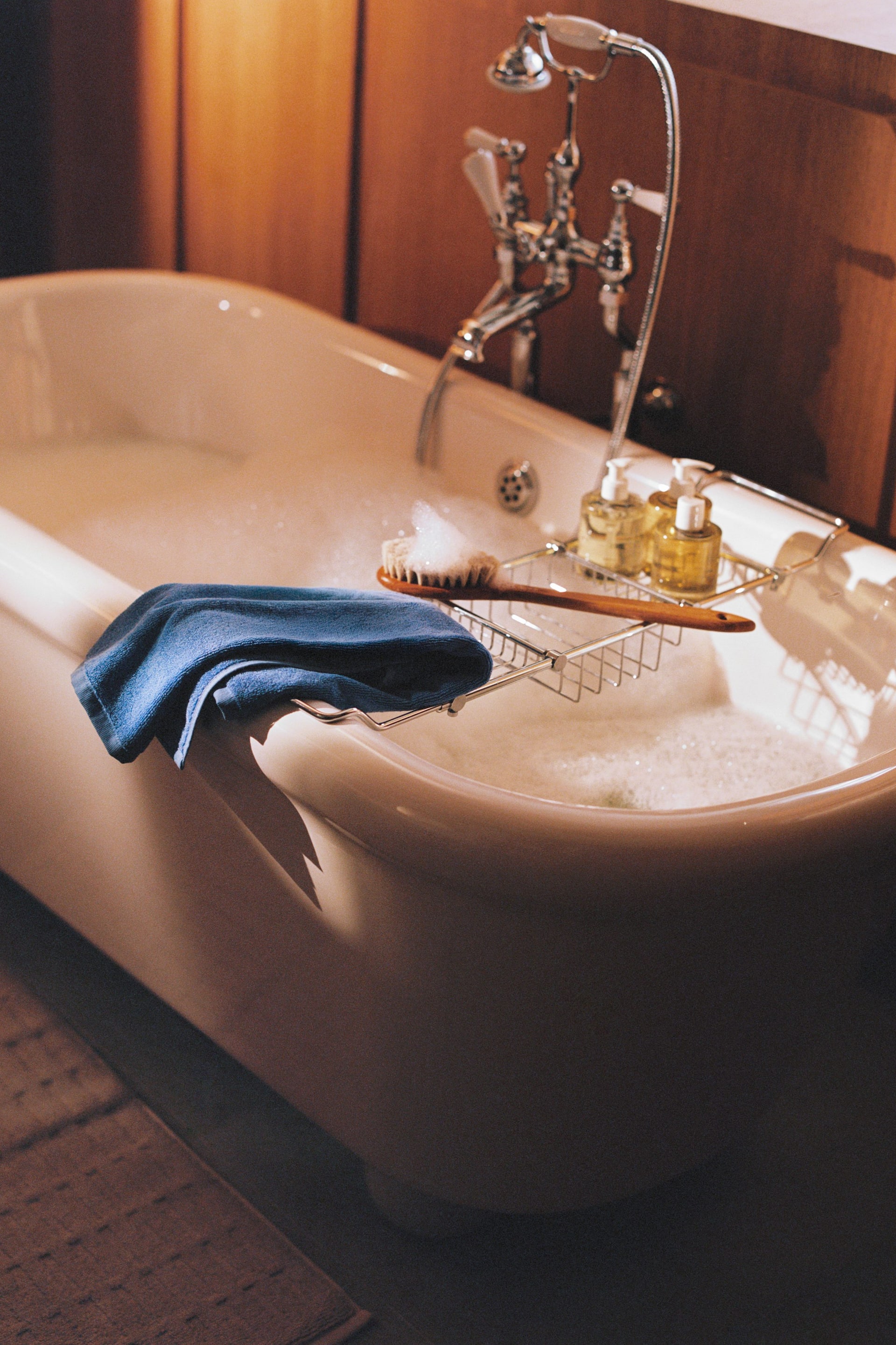 Ingrandisci l'immagine: Una vasca da bagno bianca autoportante è riempita con acqua schiumosa, dotata di un gruppo rubinetto cromato con doccetta a mano. Un vassoio cromato è appoggiato sul bordo della vasca, contenente due flaconi con dosatore e una spazzola per il corpo in legno, con un asciugamano blu piegato appoggiato accanto.