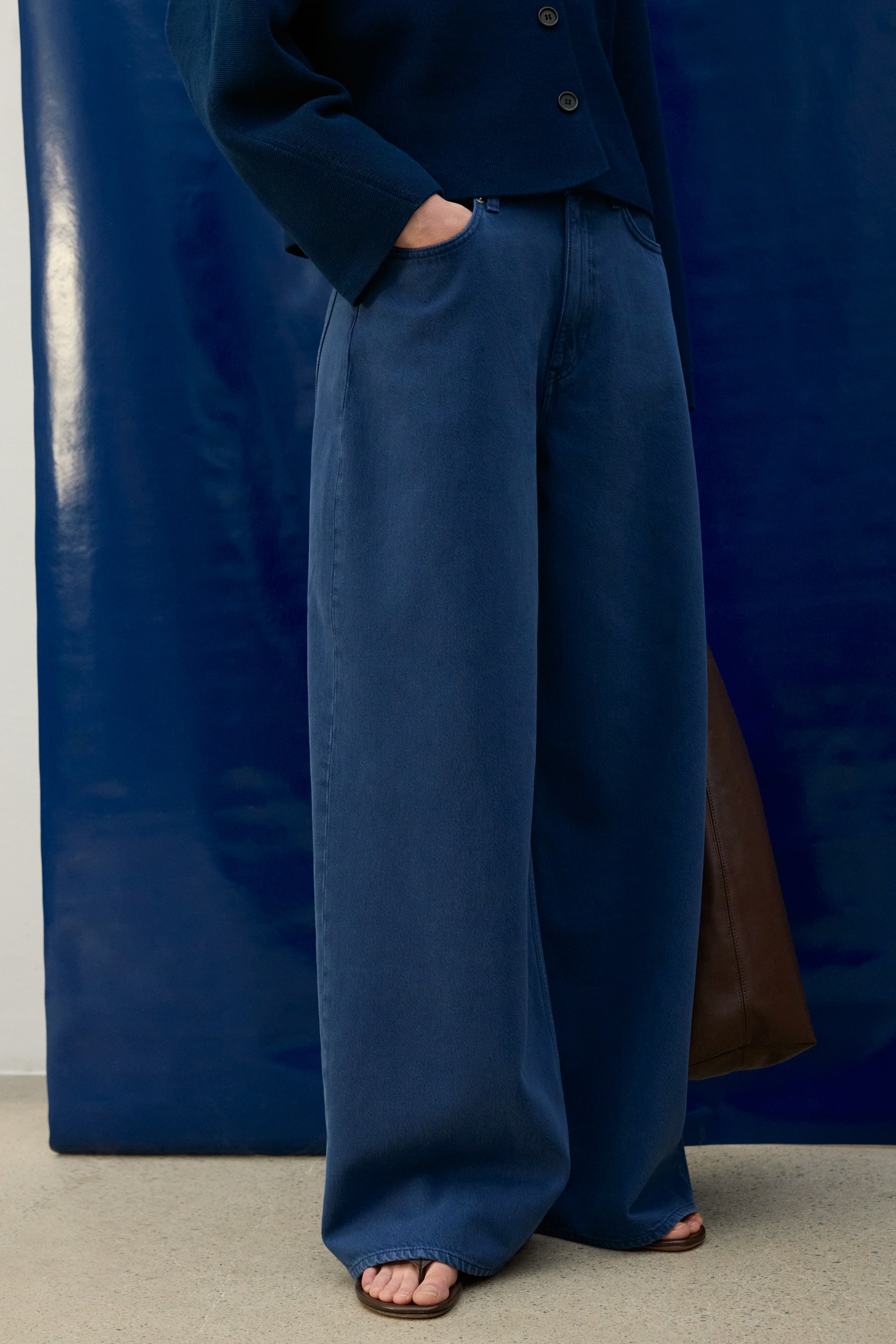 View larger image: Blue wide-leg trousers are displayed front-facing, showcasing a high-waisted design with visible front pockets. Their loose, relaxed fit flows downwards, with the full-length fabric softly pooling at the hem.