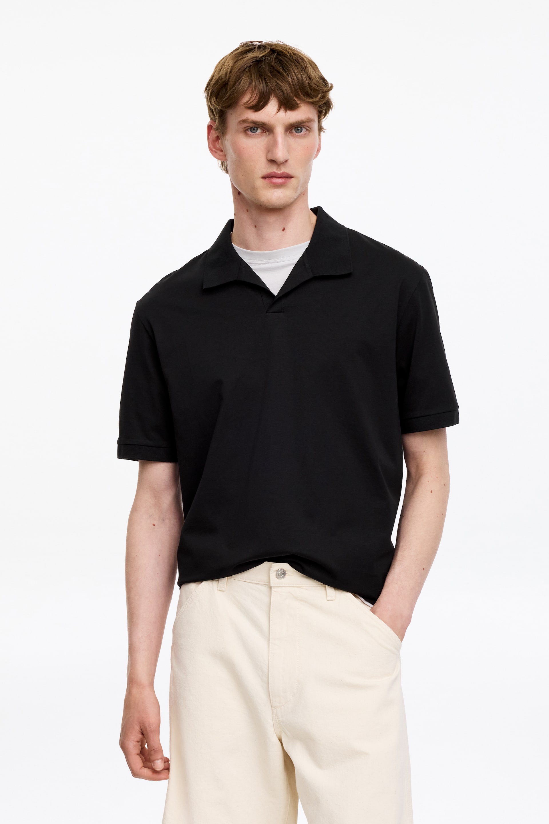 Größeres Bild anzeigen: Ein schwarzes Poloshirt wird von vorne präsentiert, mit einem offenen Kragen und kurzen Ärmeln. Es wird über einem hellgrauen T-Shirt getragen, das am Halsausschnitt sichtbar ist.