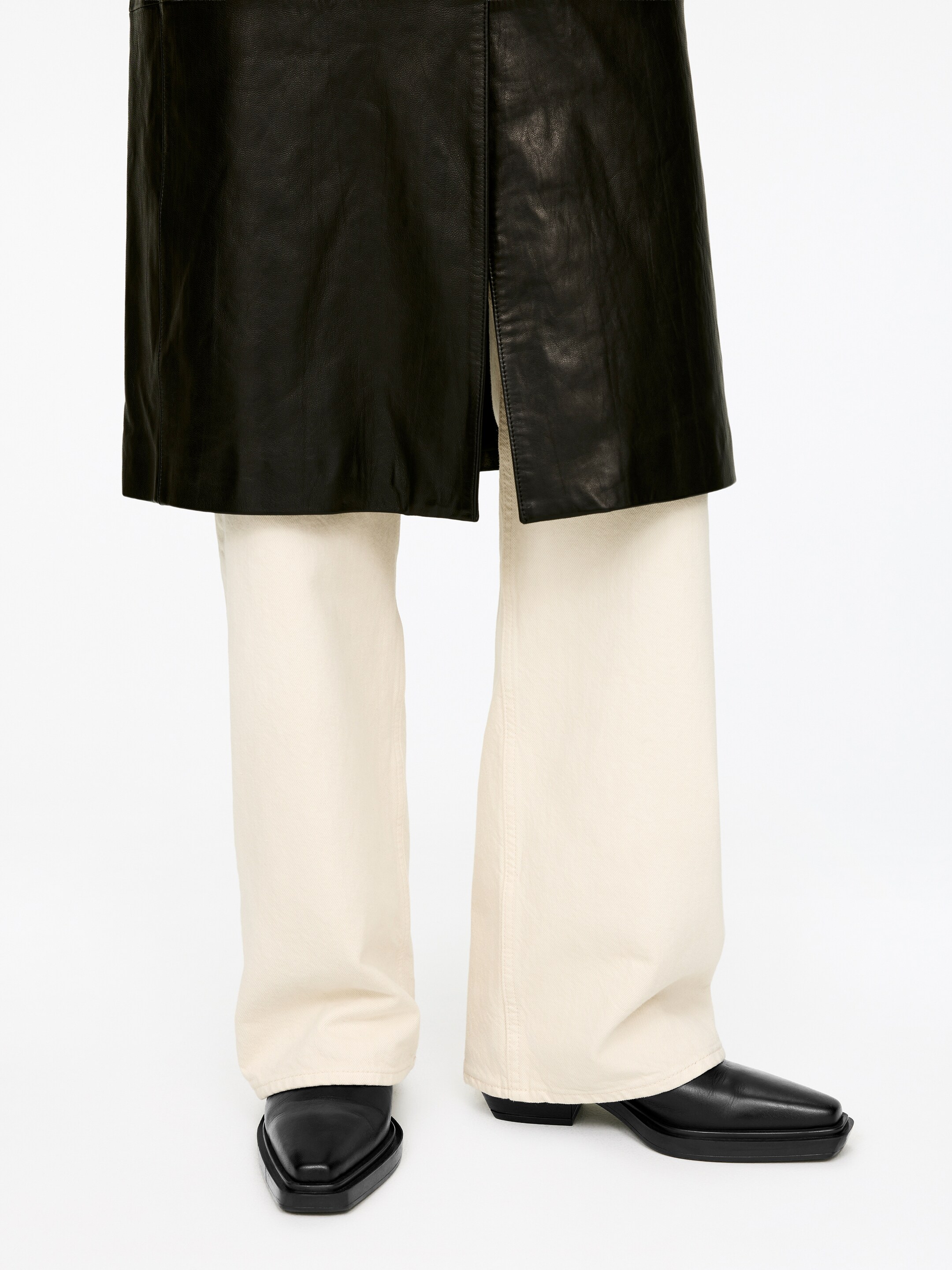 Agrandir l'image: Un pantalon large crème se présente de face, s'étend sur toute la longueur et se regroupe sur des chaussures à bout carré noires. Un vêtement en similicuir noir avec une fente à l'avant est superposé au pantalon, atteignant le niveau mi-cuisse.