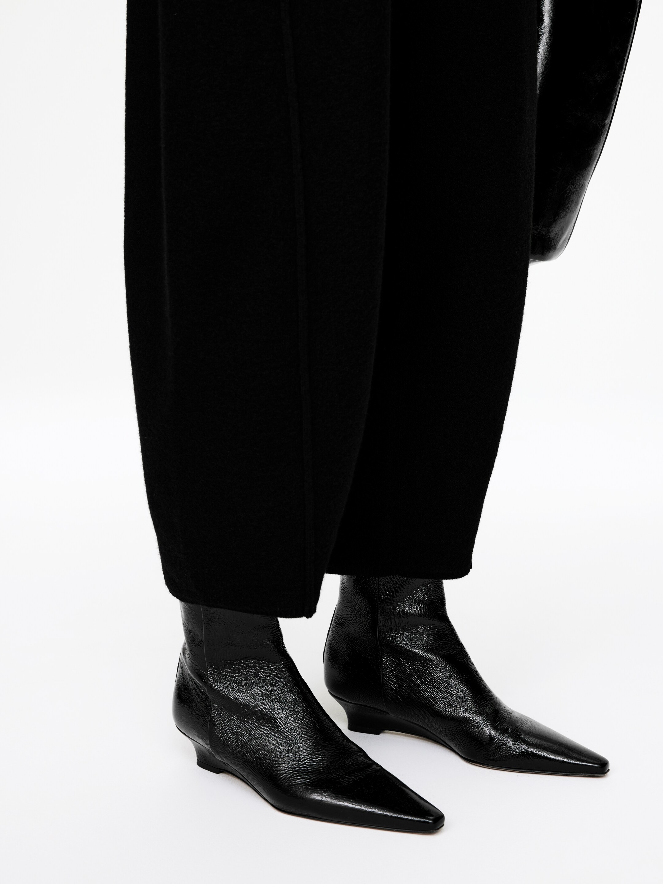 View larger image: Black ankle boots are being presented from an angled front view, featuring pointed, slightly squared toes and low, wedge-like heels. The boots are displaying a crinkled texture on the vamp and smooth ankle shafts, with black wide-leg trousers draping over them.