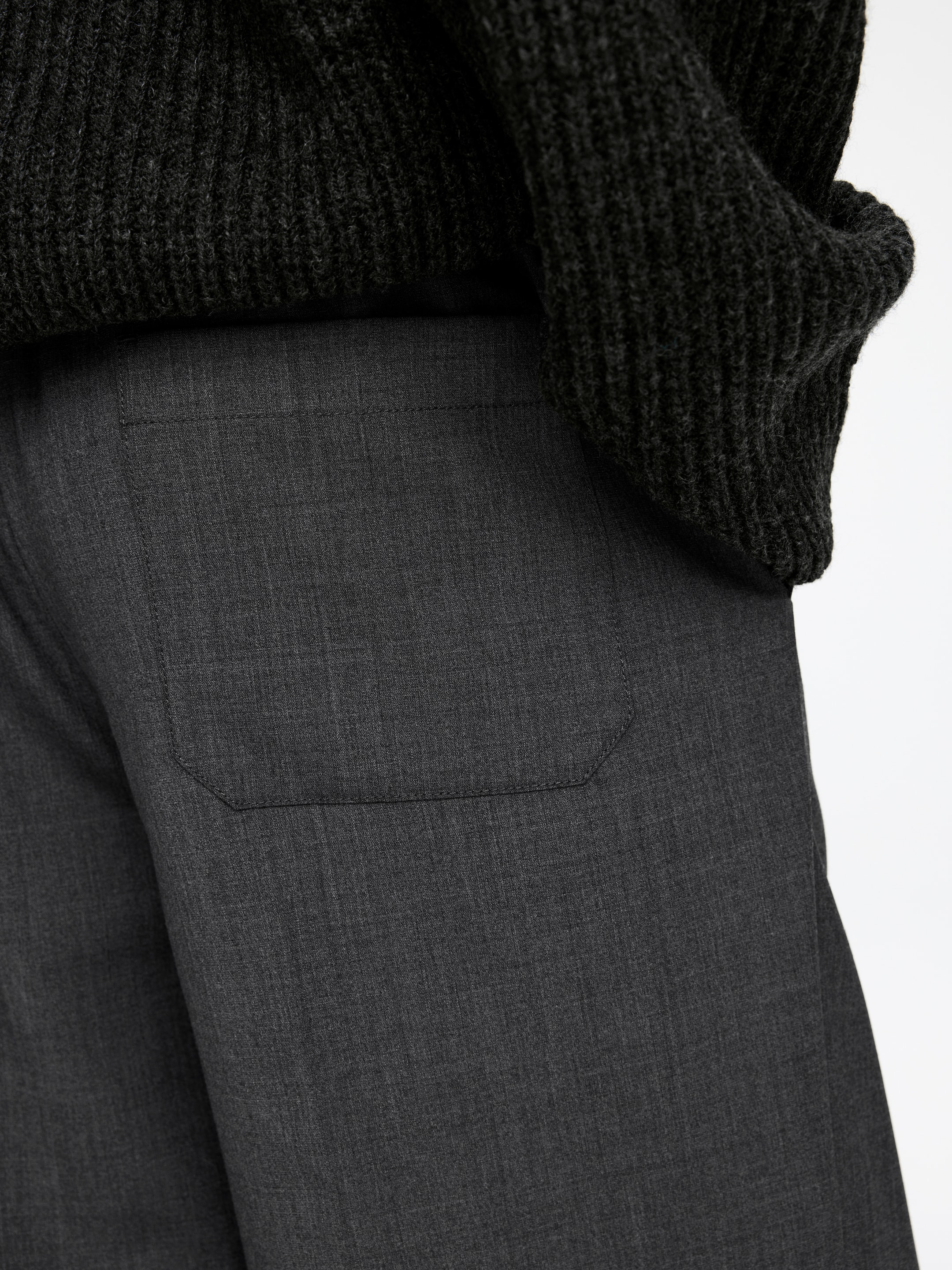 Ingrandisci l'immagine: Pantaloni grigio scuro vengono mostrati da un'angolazione laterale, caratterizzati da una sottile texture melange e una tasca a toppa rettangolare piatta con cuciture visibili lungo il bordo superiore. Un maglione scuro a maglia grossa viene indossato sopra i pantaloni, coprendo l'area della cintura.
