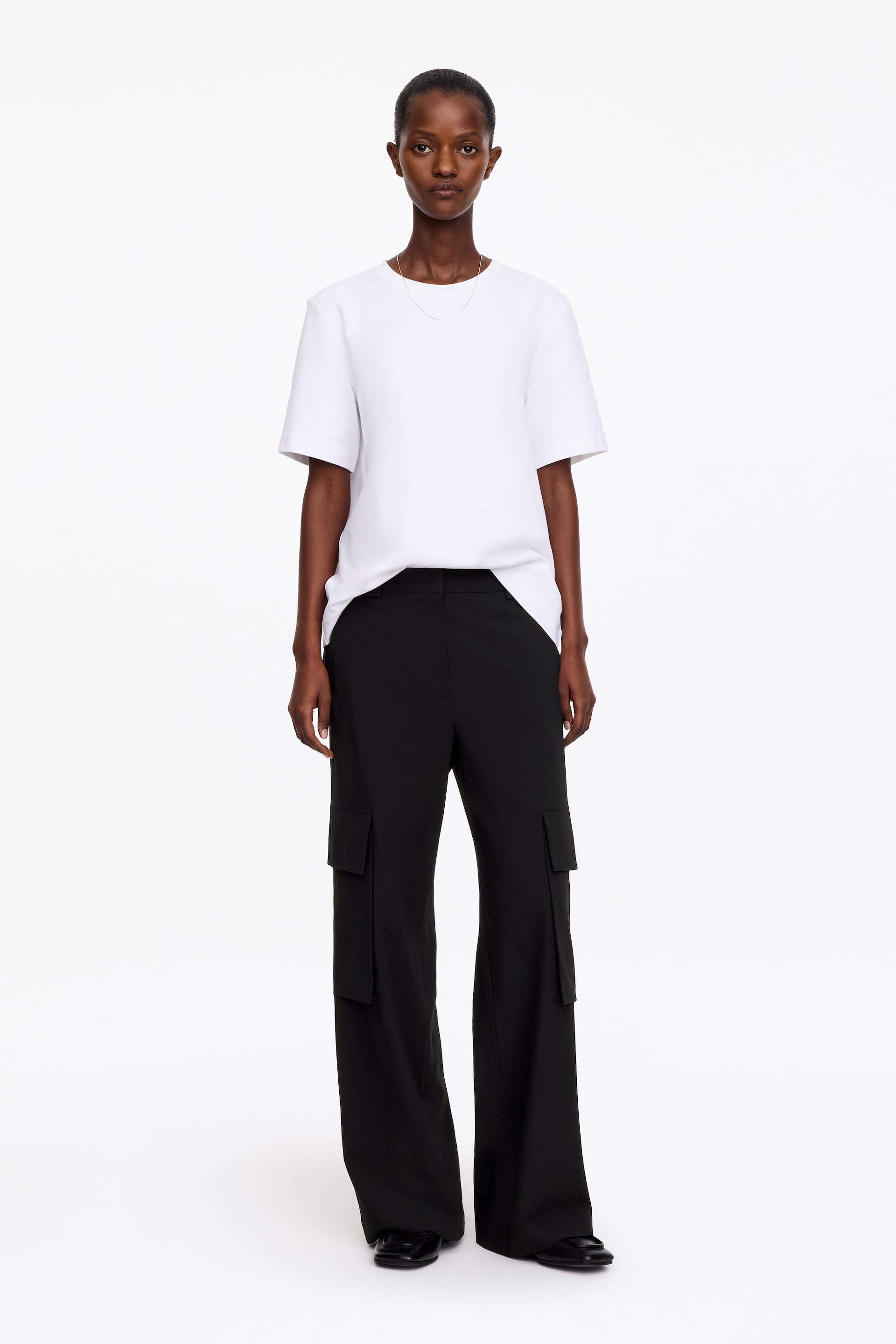 Vezi imaginea mai mare: Pantaloni negri cu croială largă, cu talie medie și buzunare cu clapetă tip cargo pe lateralul picioarelor, purtați de model. Pantalonii sunt asortați cu un tricou alb cu mâneci scurte și pantofi negri.