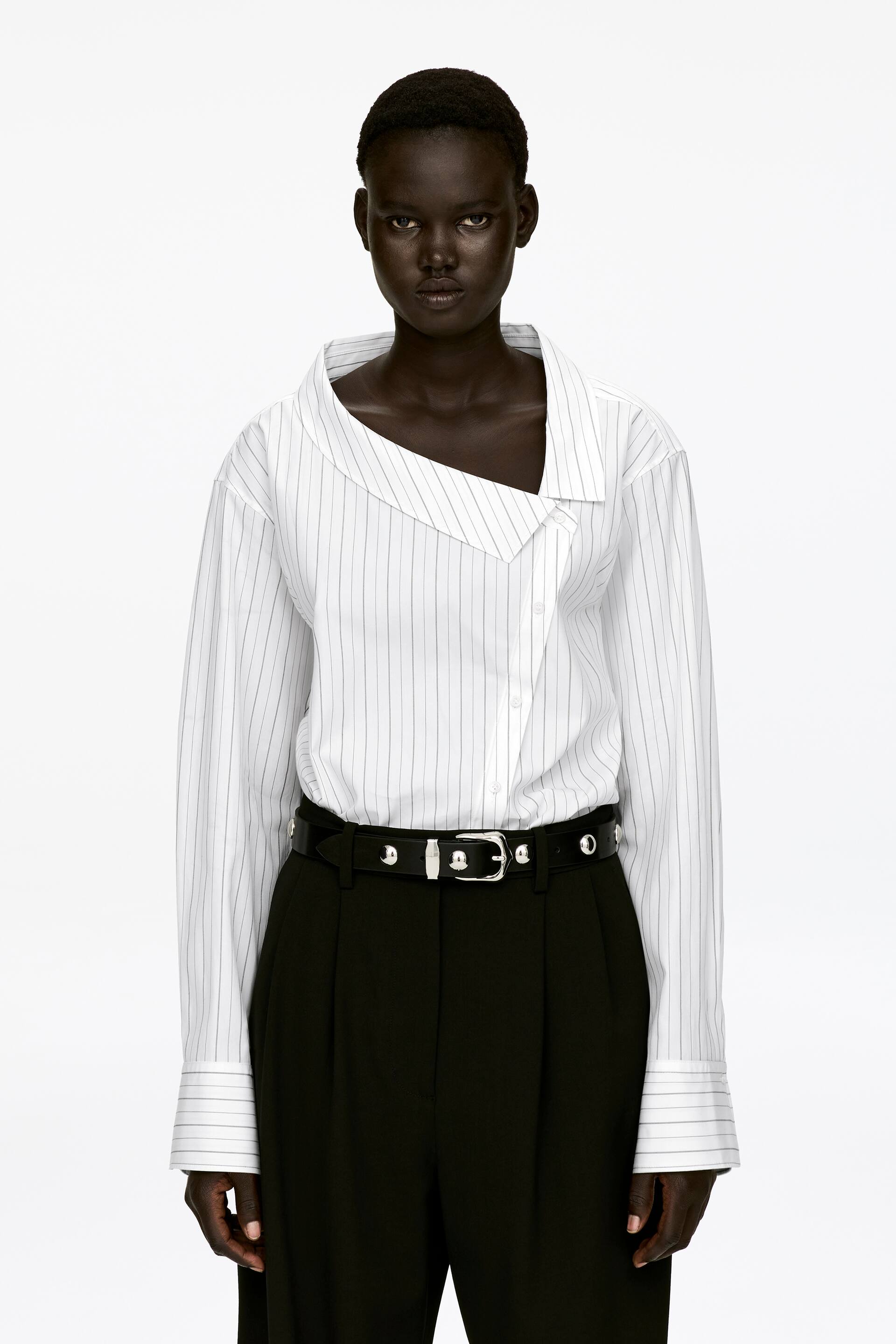 Vis større bilde: En hvit bomullsbluse med pinstripe-mønster, som har en asymmetrisk krage og diagonale knapper, bæres av modellen. Blusen er avsluttet med vide ermer med mansjetter og har en normal passform, stukket ned i svarte plissébukser og stylet med et svart belte.