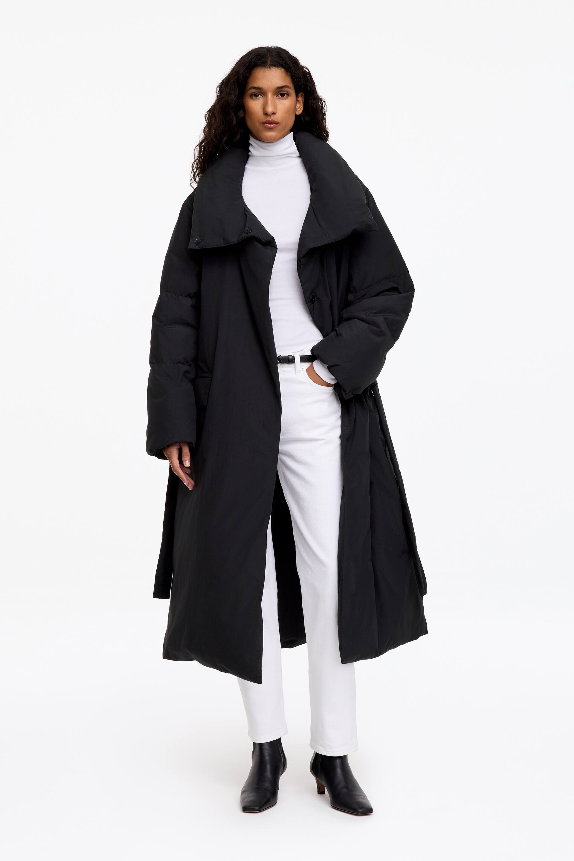View larger image: The black Re:Down puffer coat, featuring a longline silhouette and a wide, oversized collar, is worn open by the model. It is layered over a white roll-neck top and white trousers, paired with black ankle boots.