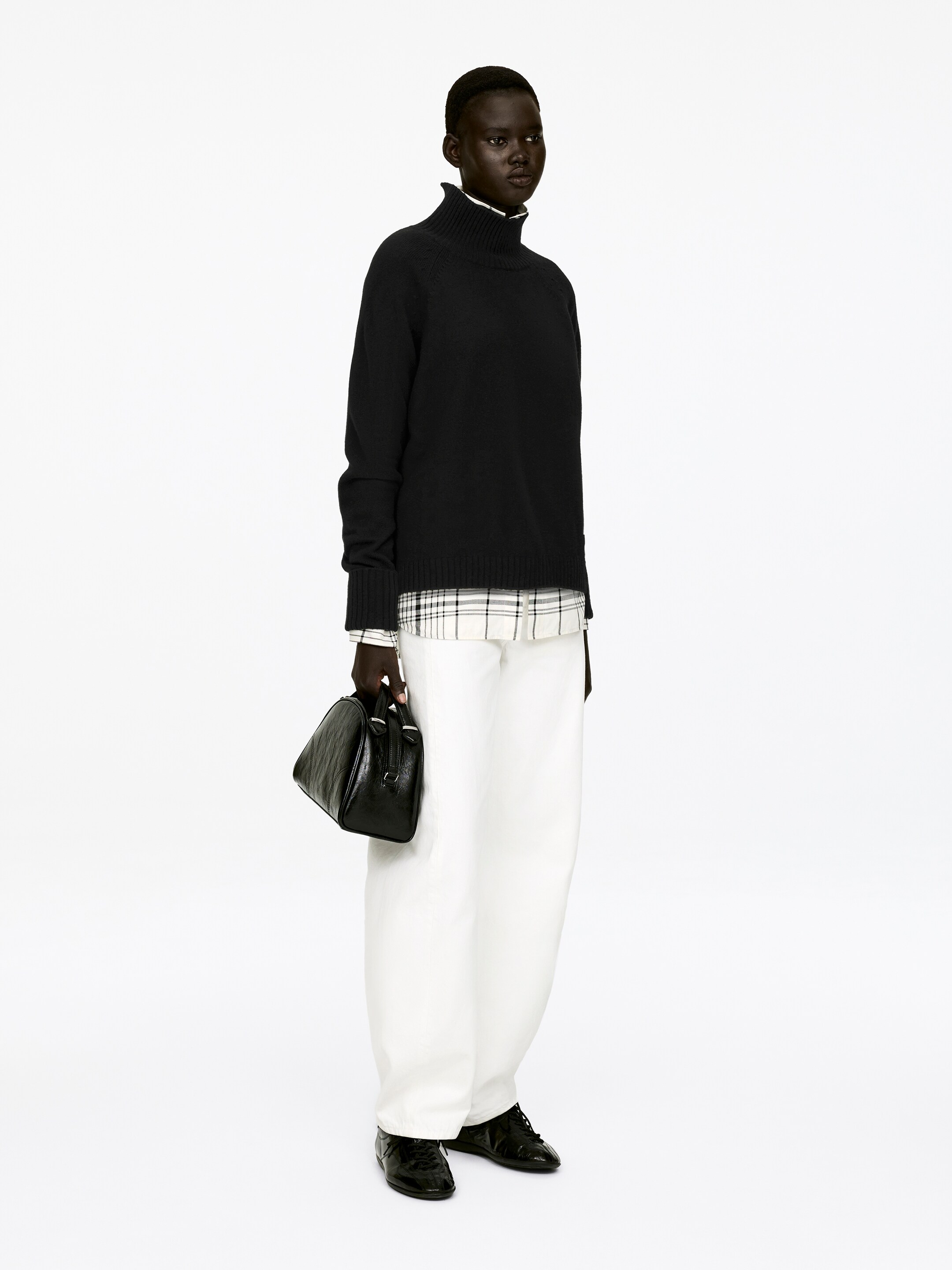 View larger image: A black regular-fit roll-neck jumper, featuring raglan sleeves and folded cuffs, is being worn by the model, with a white and black plaid shirt visibly layered underneath at the collar and hem.