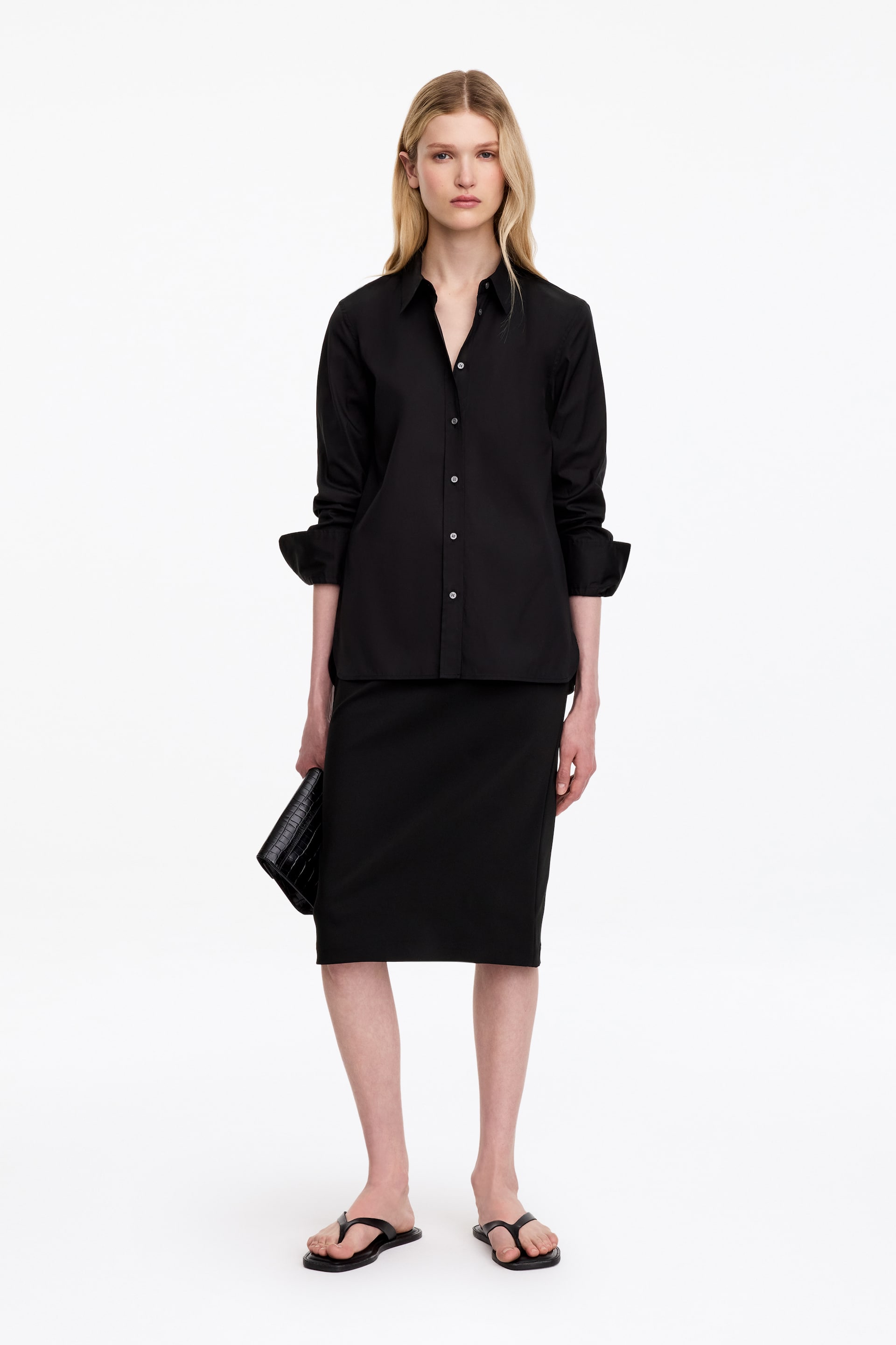 View larger image: A black slim-fit pencil skirt with a straight hem is worn by the model and falls to the knee. The skirt is styled with a matching black button-down shirt, worn untucked over the waistband.