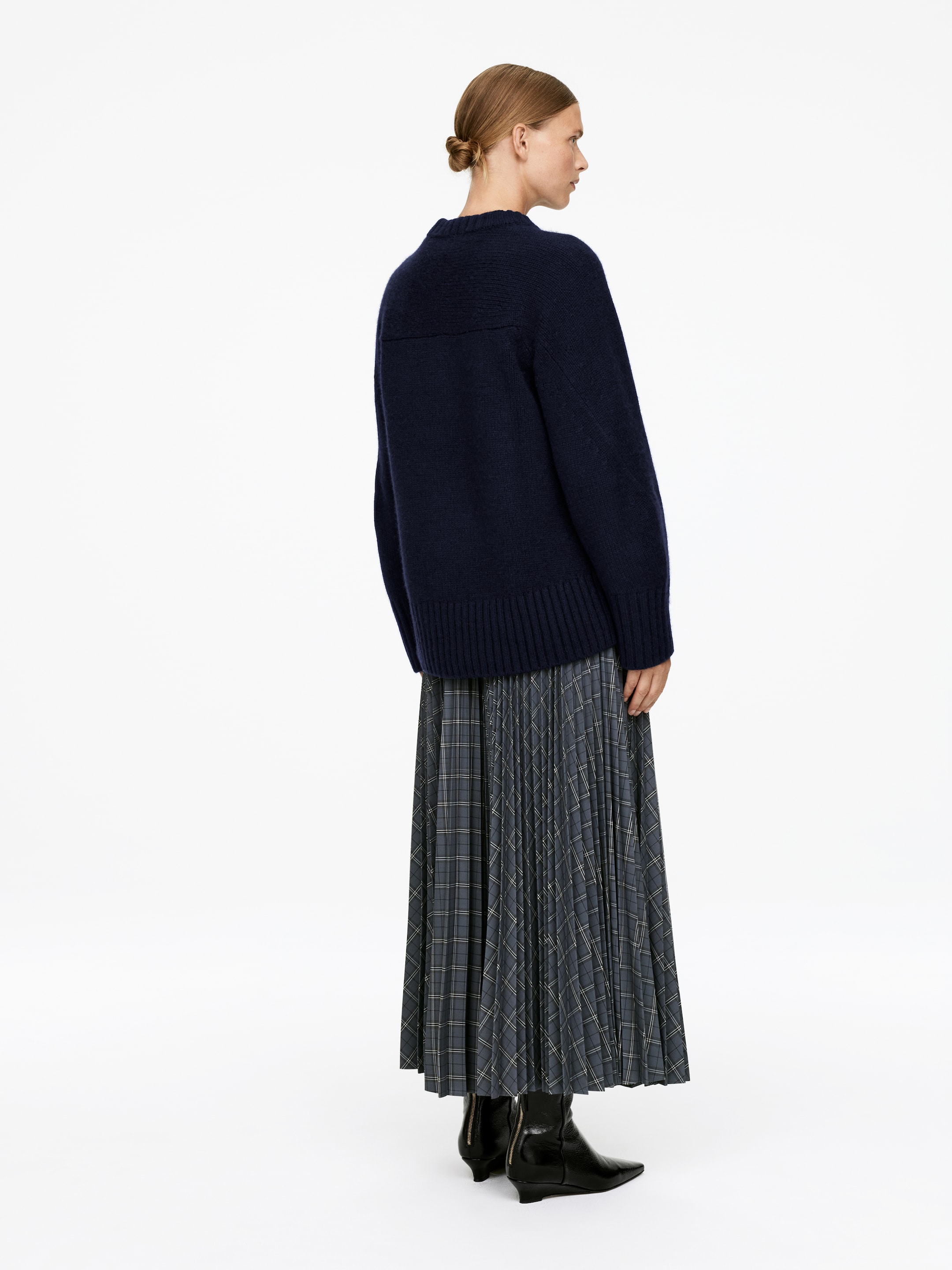 Visualizza immagine più grande: Un maglione lavorato a maglia blu navy con una vestibilità rilassata, un orlo a coste e dettagli di cuciture visibili sulla parte superiore della schiena è indossato. Una lunga gonna a pieghe a quadri in tonalità di grigio e blu è indossata sotto il maglione, estendendosi fino alle caviglie.