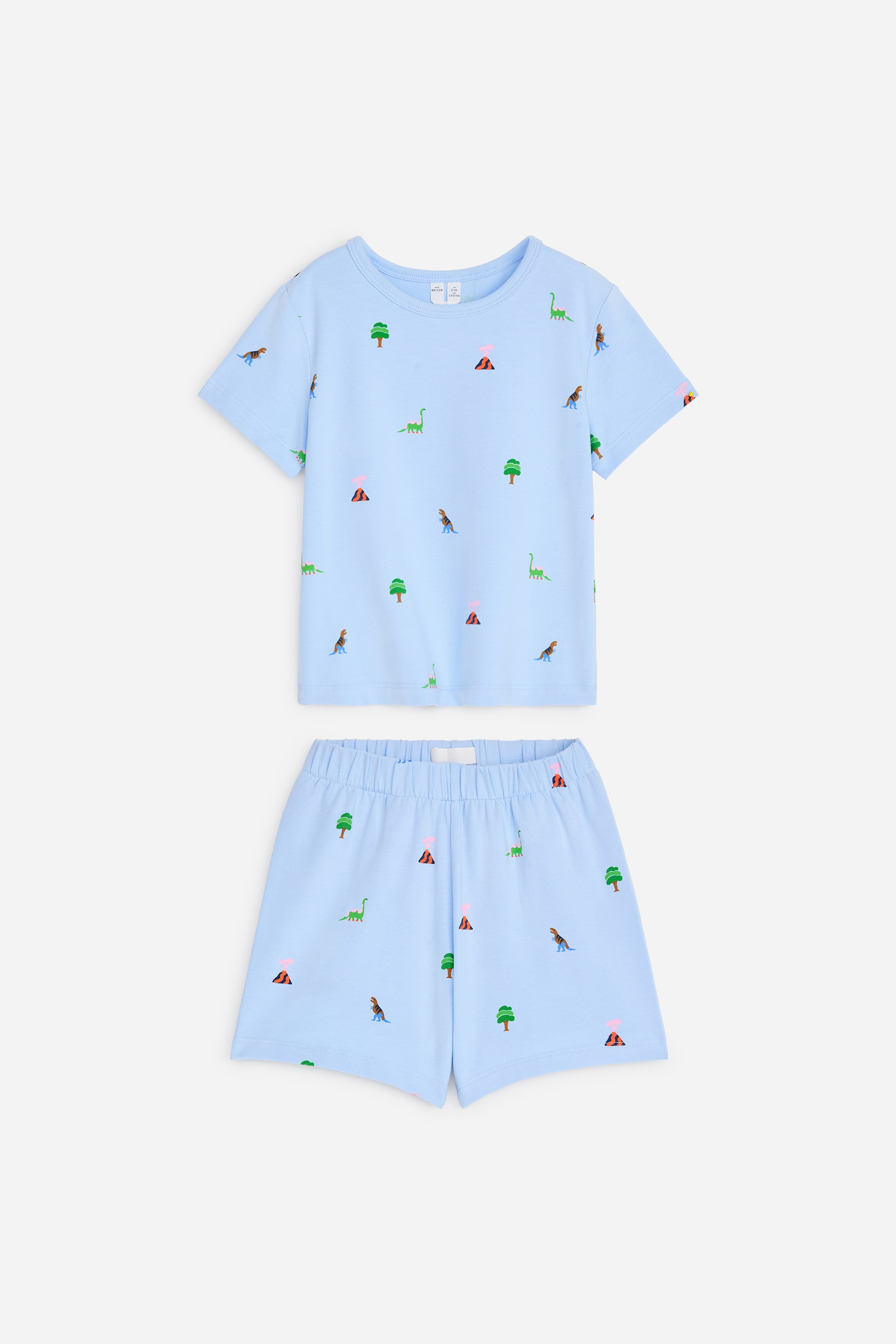 Visa större bild: Ett ljusblått pyjamasset ligger platt och är vänt framåt, bestående av en kortärmad T-shirt och matchande shorts. Båda plaggen har ett upprepat mönster av gröna dinosaurier, gröna träd och vulkaner med rosa toppar, där T-shirten har en rund hals och shortsen har en elastisk midja.