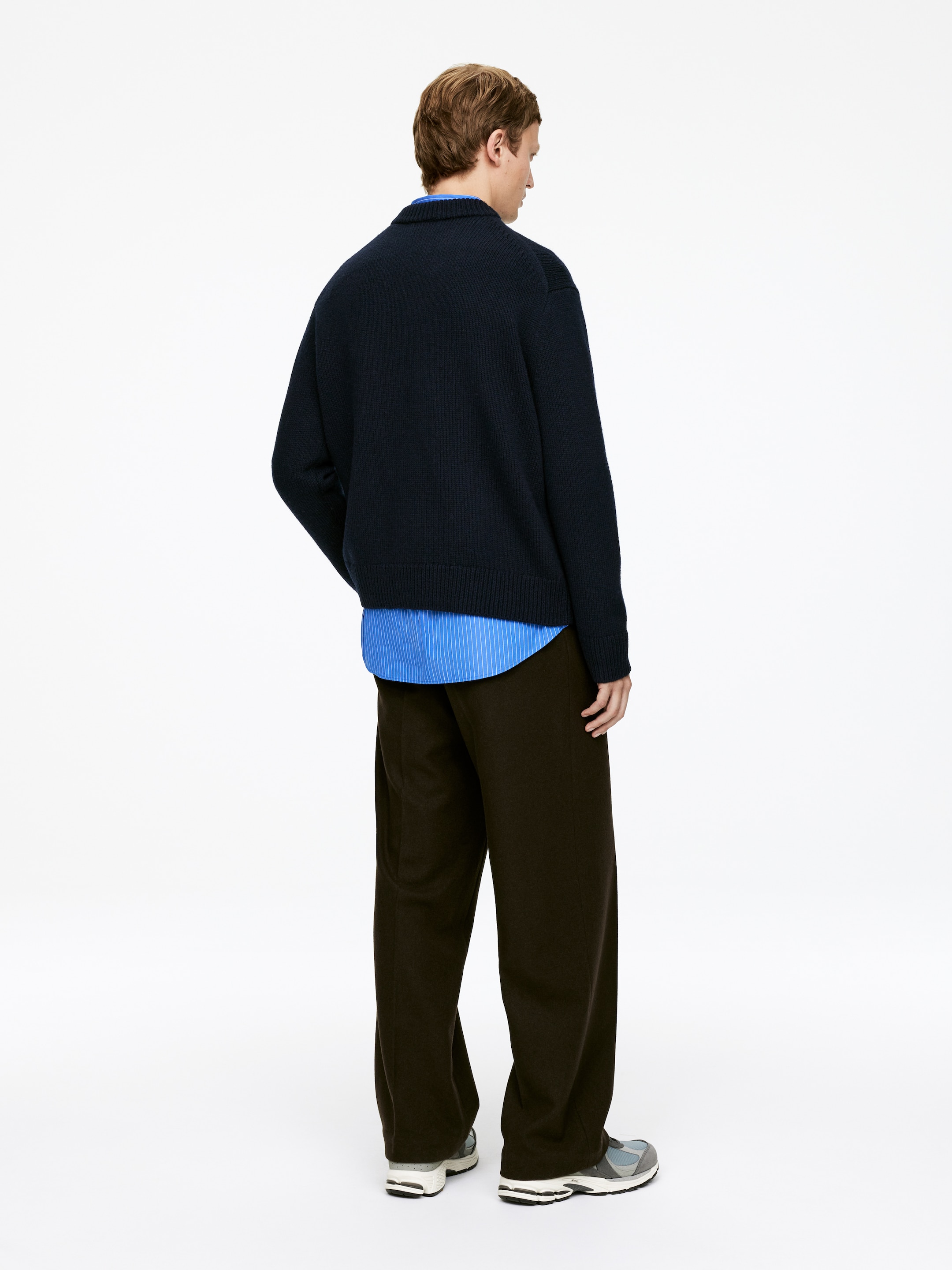 View larger image: A navy knitted collared sweater is being shown from the back, and is featuring a relaxed fit. It is being layered over a light blue striped shirt and is being worn with dark brown wide-leg trousers.