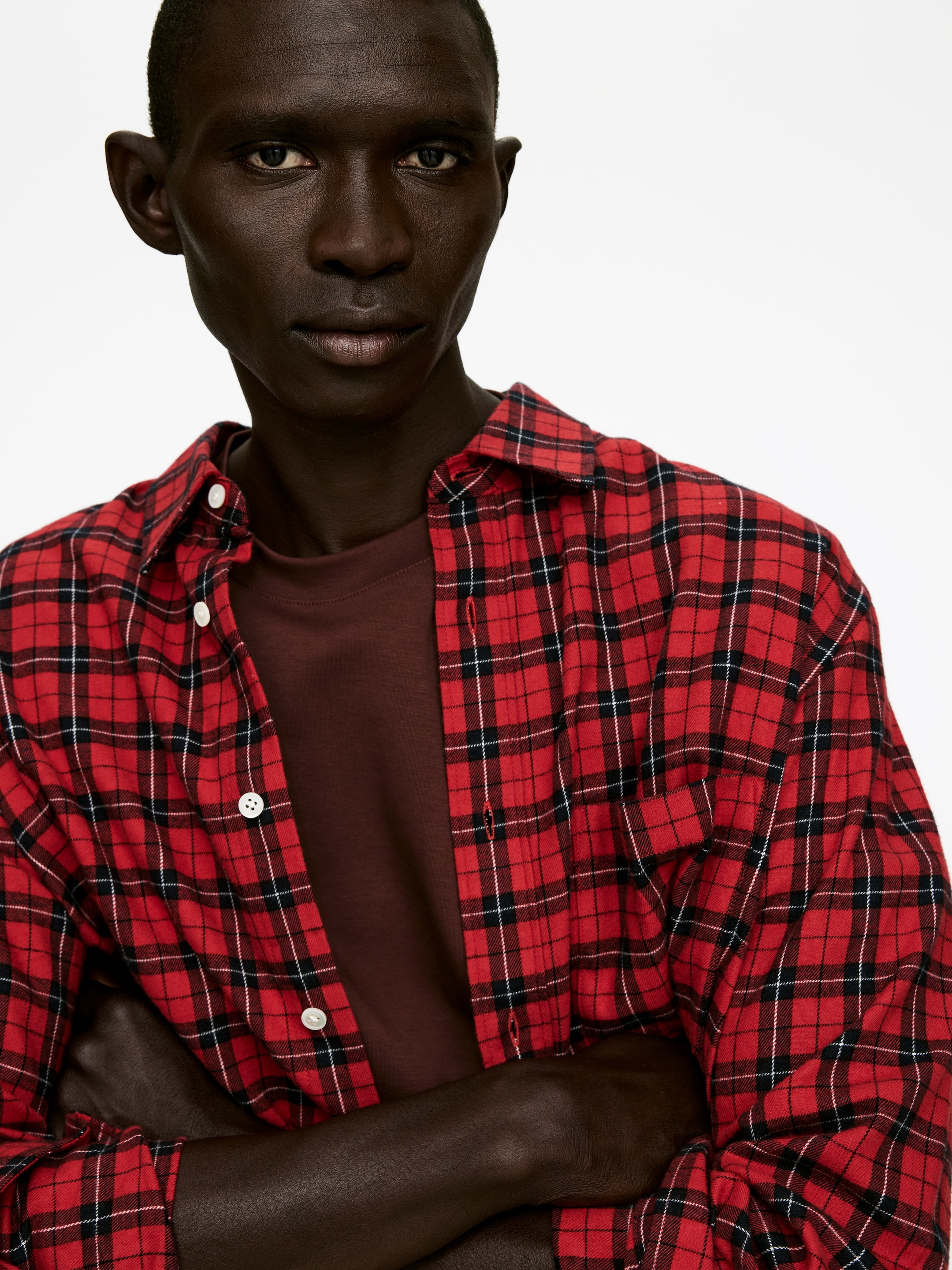 Visa större bild: En röd rutig skjorta bärs uppknäppt över en mörkbrun t-shirt. Skjortan har en röd bas med korsande svarta och tunna vita linjer som bildar ett tartanmönster, och visar vita knappar längs sin främre placket.