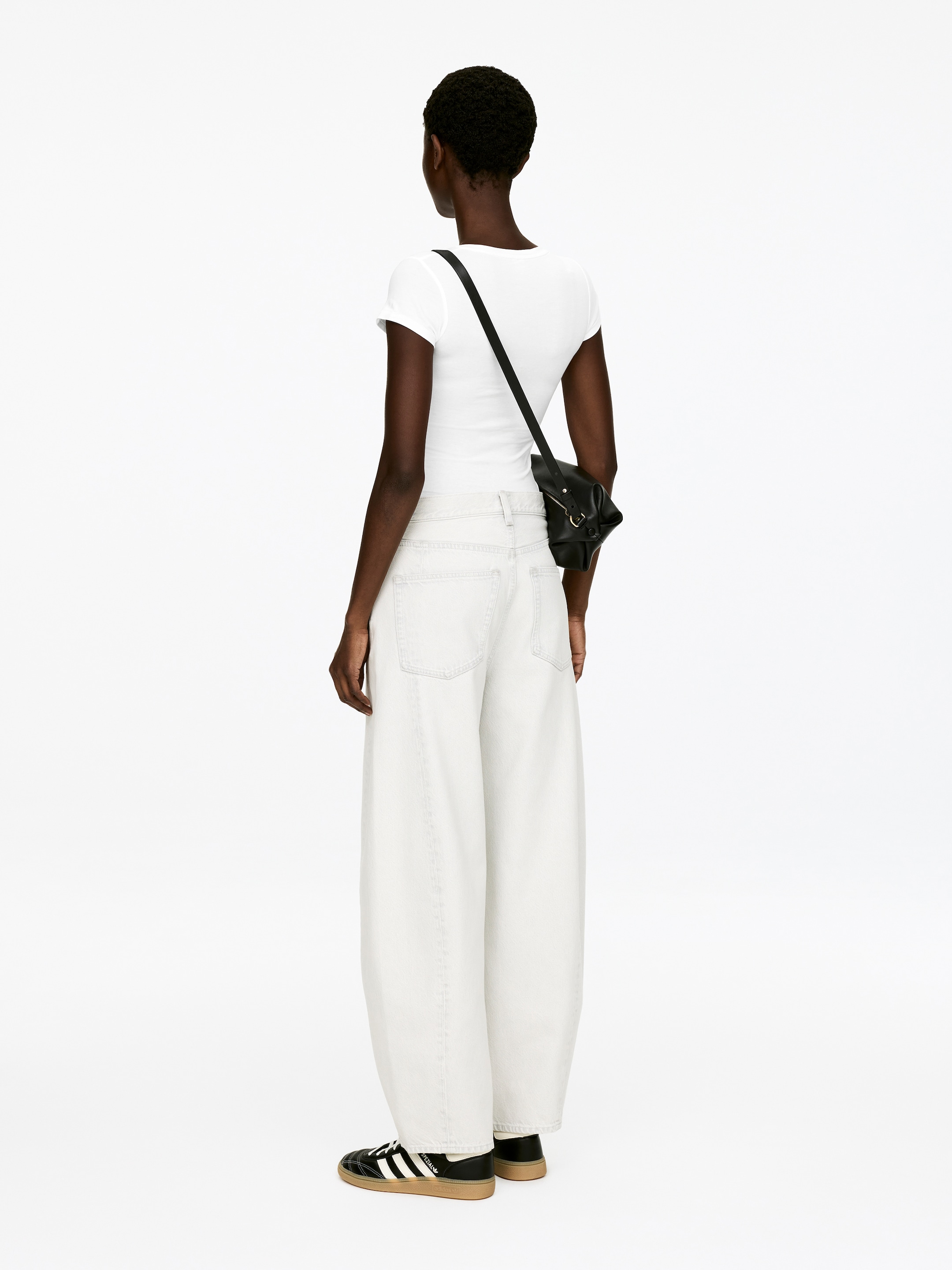 View larger image: A black crossbody bag is hanging from the shoulder, positioned at the back right hip. It is featuring a soft, gathered construction with a top flap and a prominent gold-toned metal ring detail on the front.